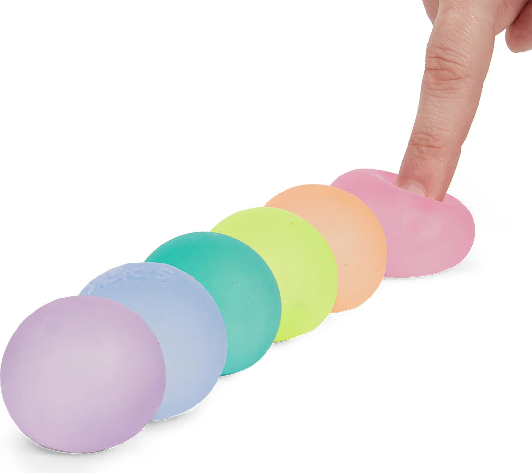A hand squeezes a pink Mini Gump from Faire's 6-pack, with the pastel office fidgets lined up neatly on a white surface.