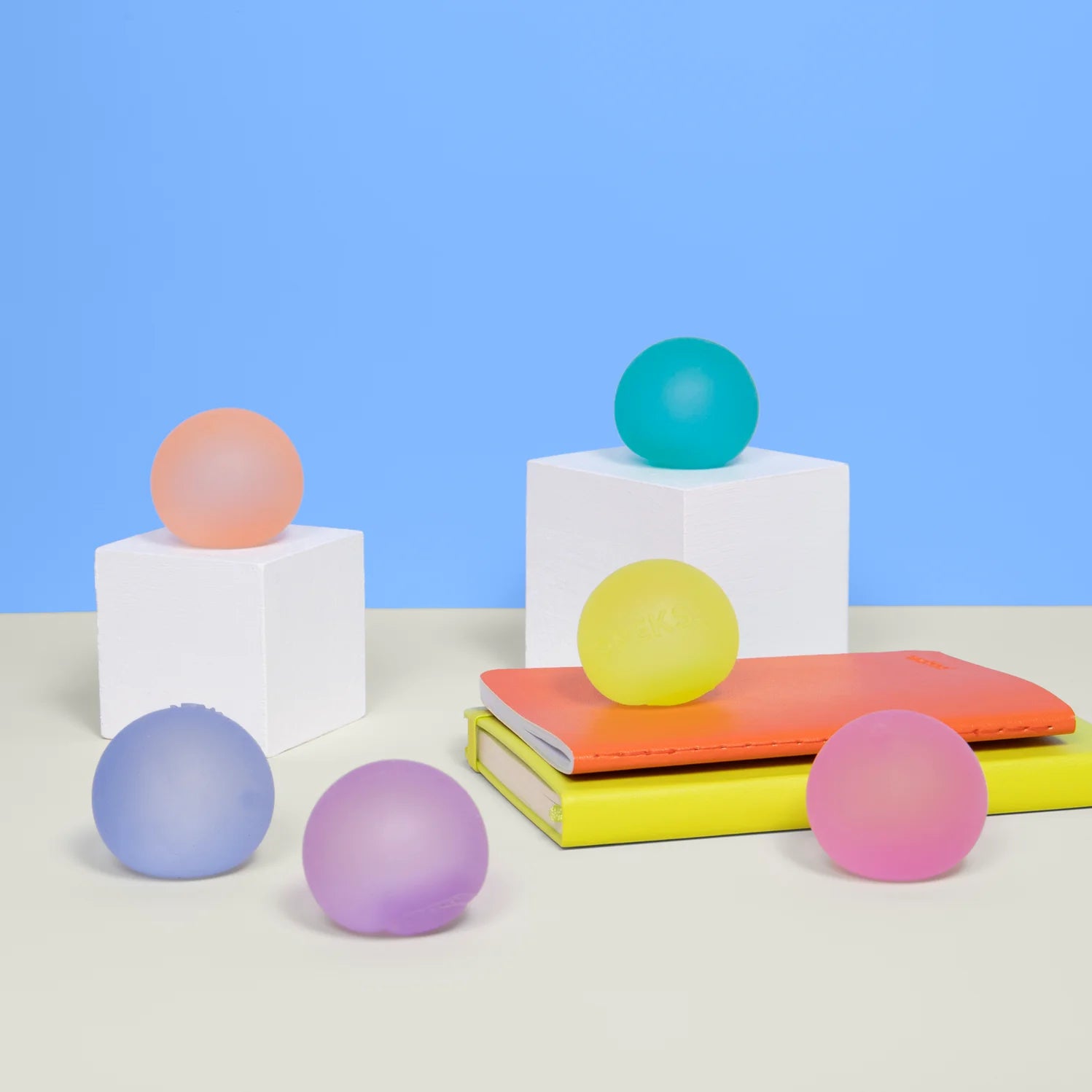 The Faire Mini Gump 6-pack features pink, orange, yellow, green, purple, and blue translucent balls on white cubes and notebooks. These playful stress balls are perfect as office fidgets against a blue and beige backdrop.