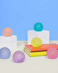 The Faire Mini Gump 6-pack features pink, orange, yellow, green, purple, and blue translucent balls on white cubes and notebooks. These playful stress balls are perfect as office fidgets against a blue and beige backdrop.
