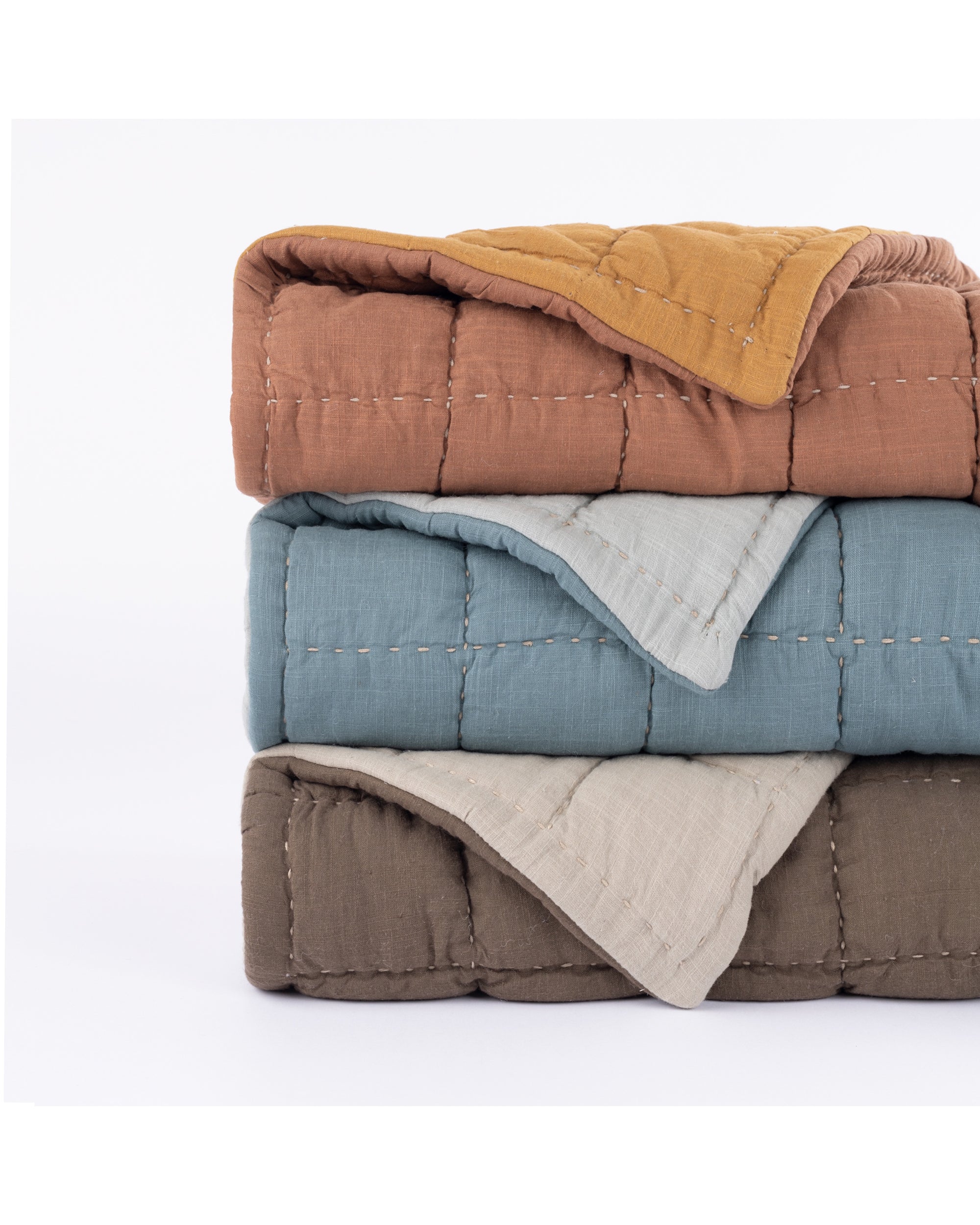 Three hand-quilted Spencer Quilts by Amity Home are stacked: the top is brown with a mustard corner, the middle is blue, and the bottom is beige and dark brown—each crafted from soft slub cotton with visible stitching.