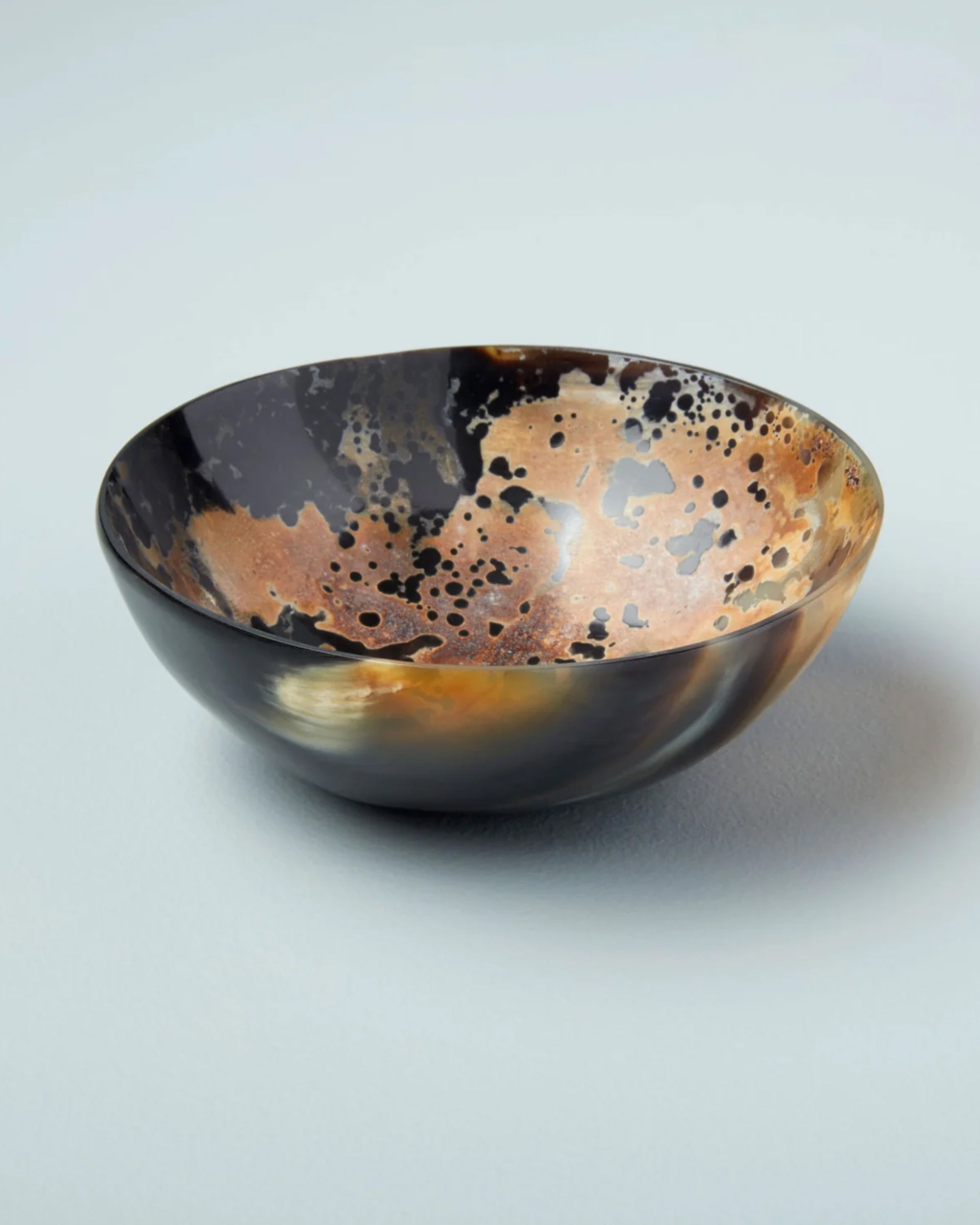 The Splatter Bowl L by Be Home is a round handmade horn bowl with a glossy, marbled pattern in black, brown, and tan that resembles natural stone. It is displayed on a light gray background.