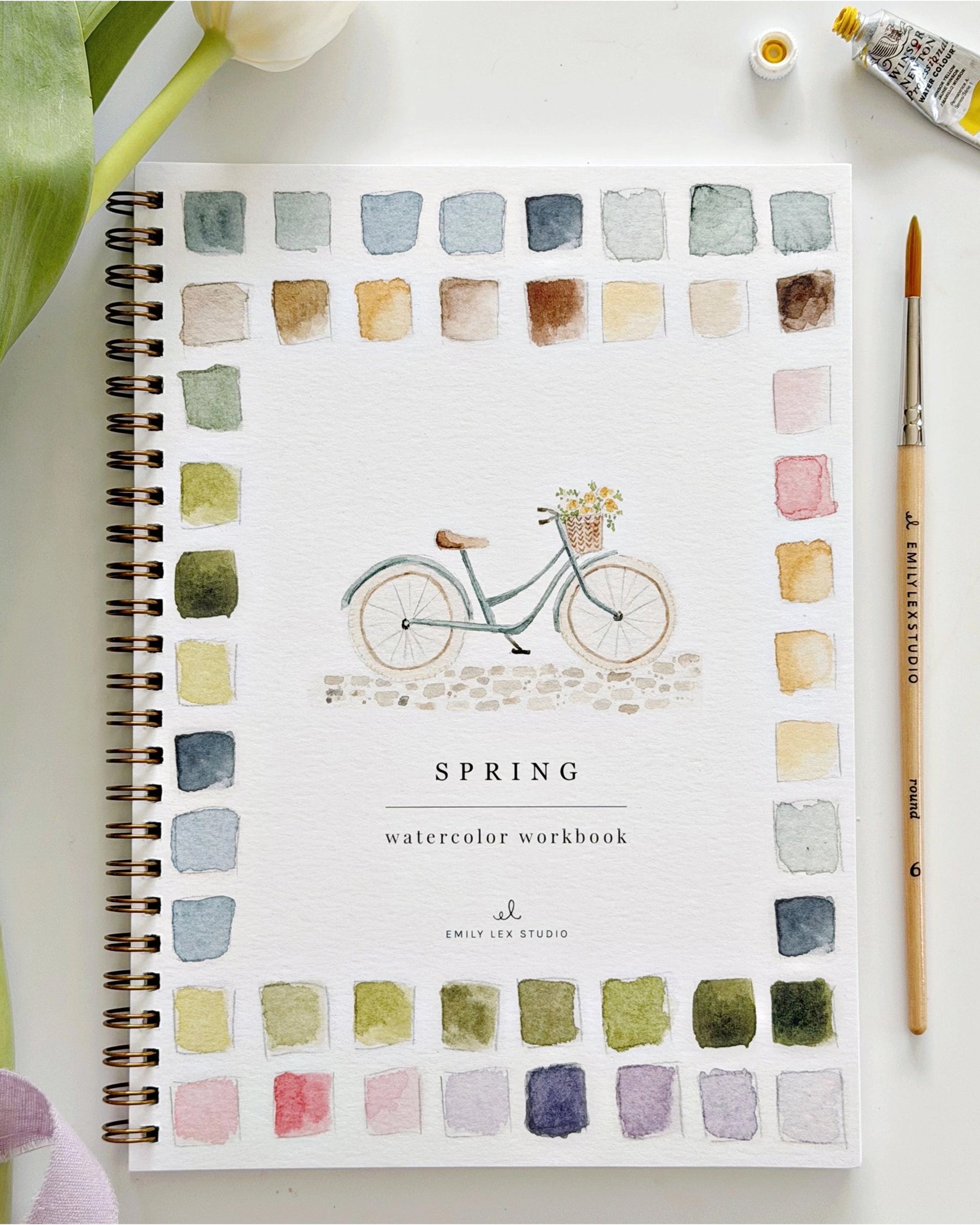 The Faire Spring watercolor workbook is spiral-bound with a bicycle cover design and colorful painted squares. Ideal for beginner art, it's shown alongside a paintbrush, paint tube, and white tulip next to the notebook.