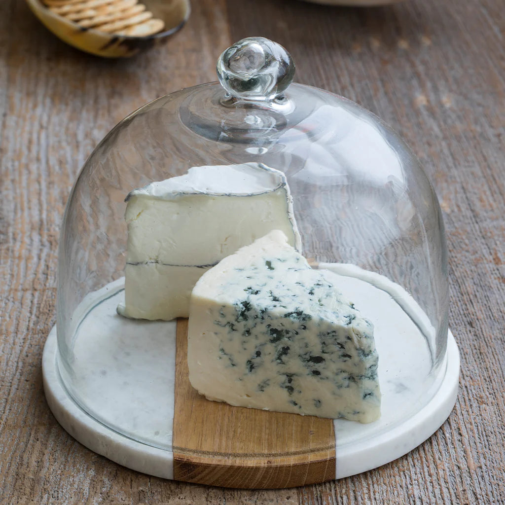 Two wedges of cheese, one blue-veined and one white, sit on a rustic table beneath the Be Home White Marble Wood Glass Cloche, featuring a handcrafted acacia board, glass dome, and marble base.
