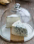 Two wedges of cheese, one blue-veined and one white, sit on a rustic table beneath the Be Home White Marble Wood Glass Cloche, featuring a handcrafted acacia board, glass dome, and marble base.