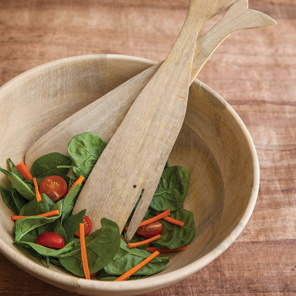 The Be Home Sydney Whale Serving Set features a handcrafted mango wood bowl with spinach, cherry tomatoes, and carrot sticks, plus two fish tail-shaped wooden salad servers on a wooden surface.