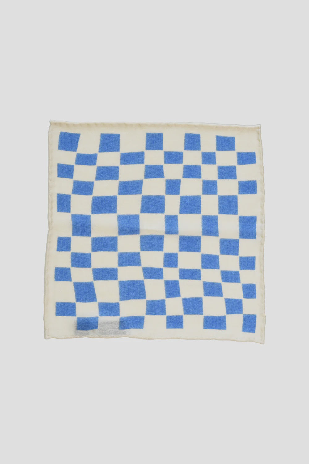 Introducing the Curated Basics MENS Blue Wool Pocket Square. This exquisite accessory features a dynamic pattern of slightly wavy blue checkered squares on a white fabric, silk-screened for a refined appearance against its plain light gray background.