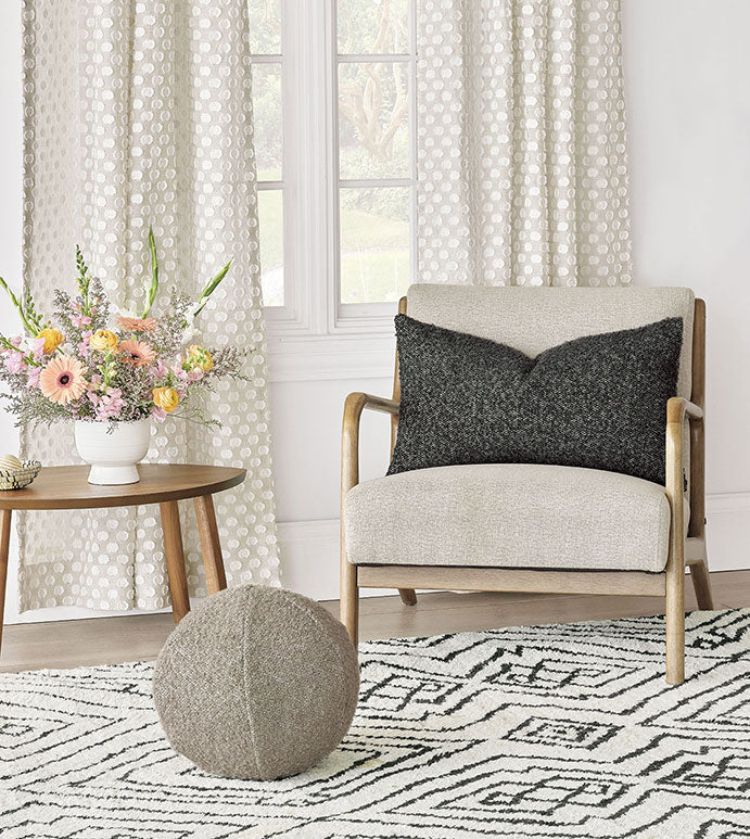 A cozy living room corner features the Eastern Accents Marl Deco Charcoal pillow on a modern armchair, alongside a geometric rug, round pouf, wooden side table, floral arrangement, and tall windows with patterned white curtains.