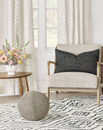 A cozy living room corner features the Eastern Accents Marl Deco Charcoal pillow on a modern armchair, alongside a geometric rug, round pouf, wooden side table, floral arrangement, and tall windows with patterned white curtains.