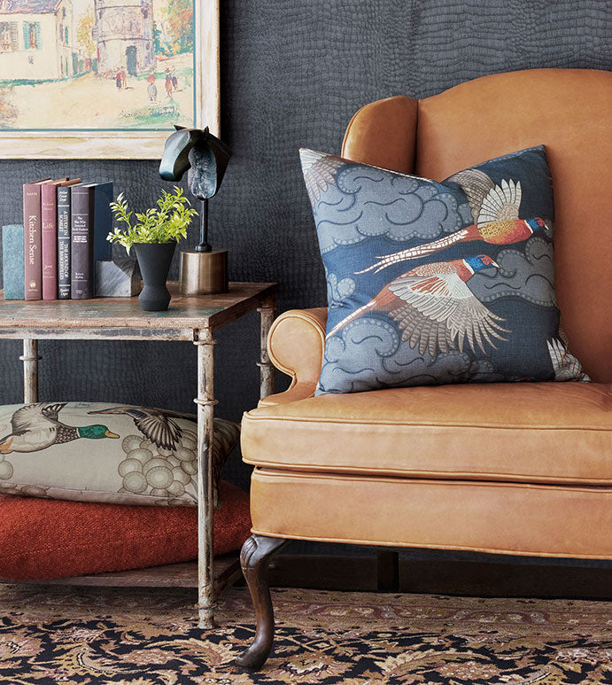 A tan leather armchair with a Barrett Aves Printed pillow by Eastern Accents sits by a rustic side table with books, a plant, and a lamp. Patterned floor cushions and another Barrett Aves Printed pillow complete the cozy scene.