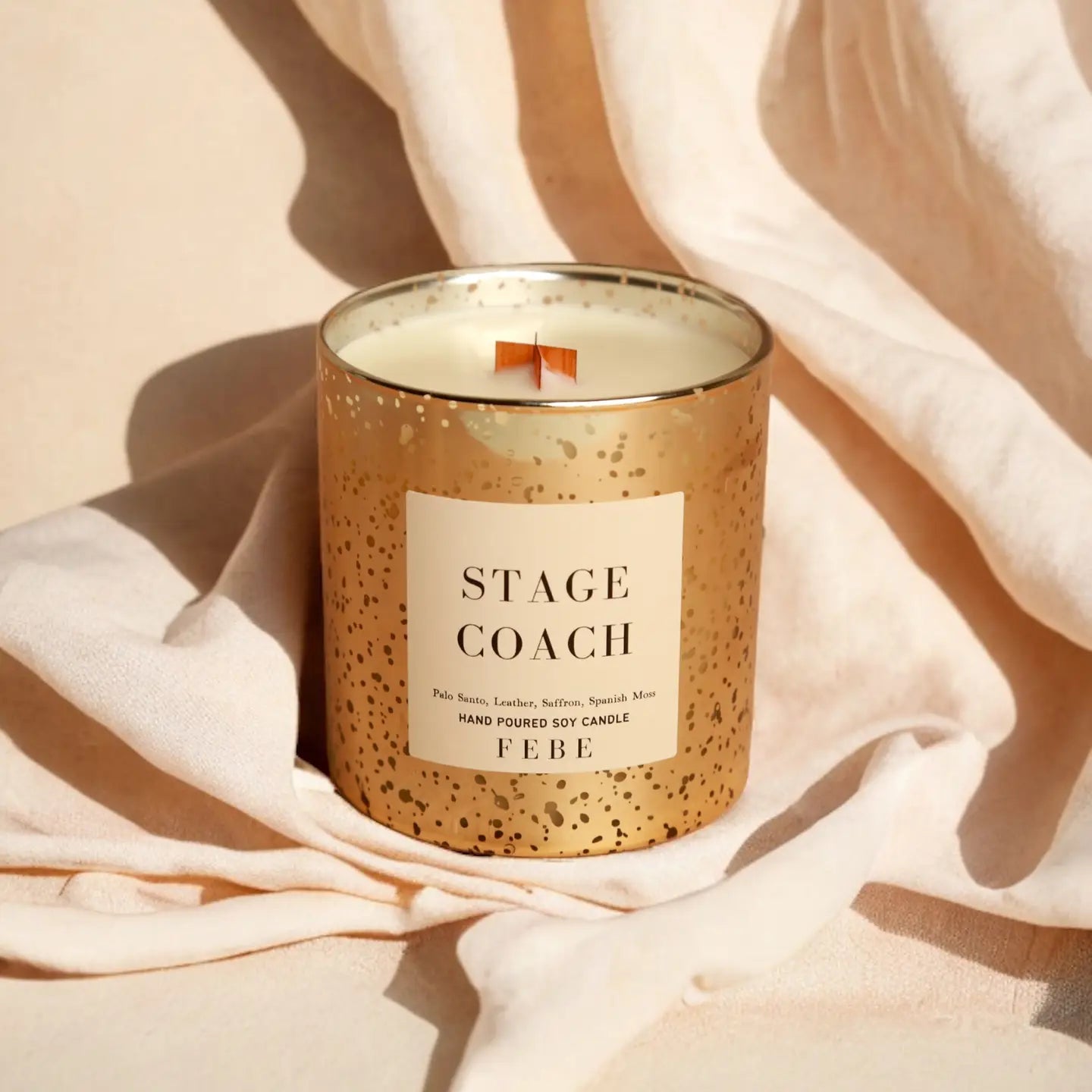 A scented candle labeled &quot;FEBE candle by Faire&quot; in a speckled gold container with a wooden wick, set against a soft beige Bungalow-style fabric background.