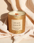 A scented candle labeled "FEBE candle by Faire" in a speckled gold container with a wooden wick, set against a soft beige Bungalow-style fabric background.