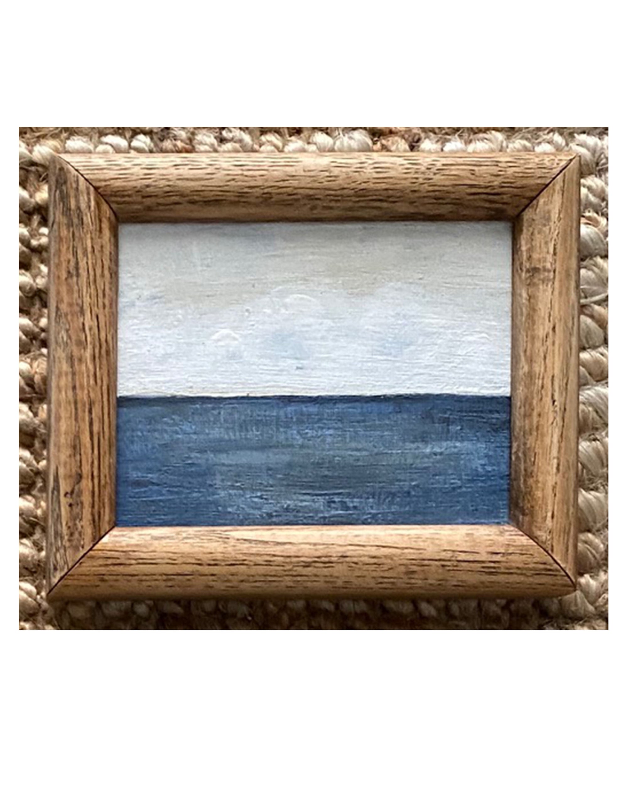 SKY & STARS by Tak Design is a small, framed artwork featuring a minimalist seascape—blue ocean under a pale sky—in a vintage frame displayed on woven fabric.