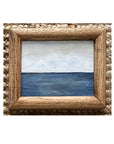 SKY & STARS by Tak Design is a small, framed artwork featuring a minimalist seascape—blue ocean under a pale sky—in a vintage frame displayed on woven fabric.
