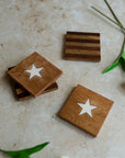 Four Faire Stars & Stripes Coasters made of mango wood rest on a beige surface: two with white star inlays, one striped, and one partially hidden. Green plant stems lend a fresh farmhouse decor accent to the scene.