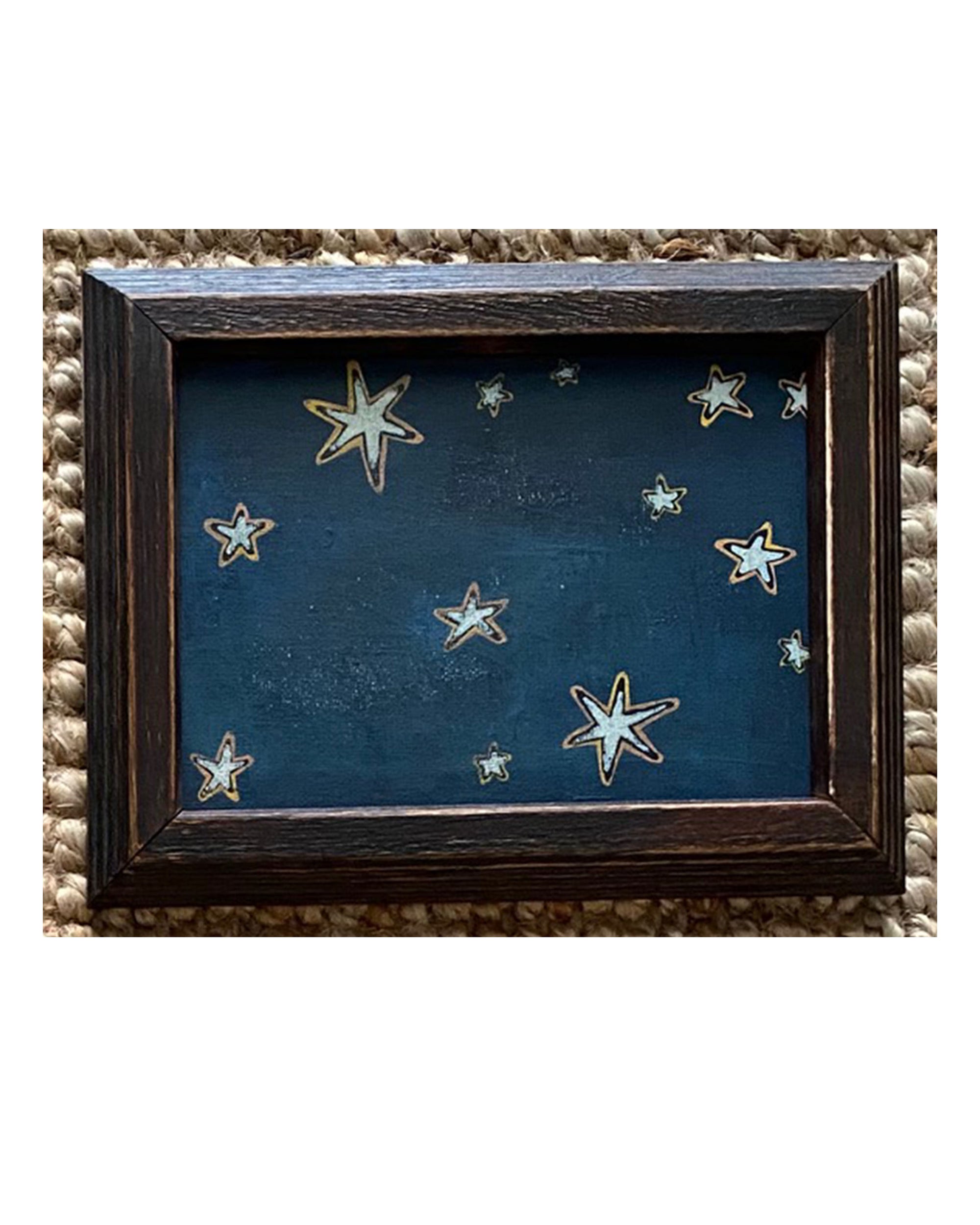 The SKY & STARS by Tak Design is a small dark vintage frame featuring hand-painted white and yellow stars on a deep blue background, all set against a textured beige surface.