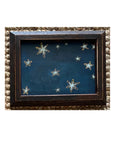 The SKY & STARS by Tak Design is a small dark vintage frame featuring hand-painted white and yellow stars on a deep blue background, all set against a textured beige surface.