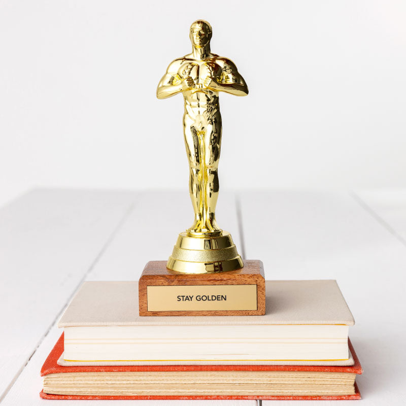 A JE Trophy styled like a human figure stands atop a stack of books with a plaque reading &quot;STAY GOLDEN&quot; on a white background.
