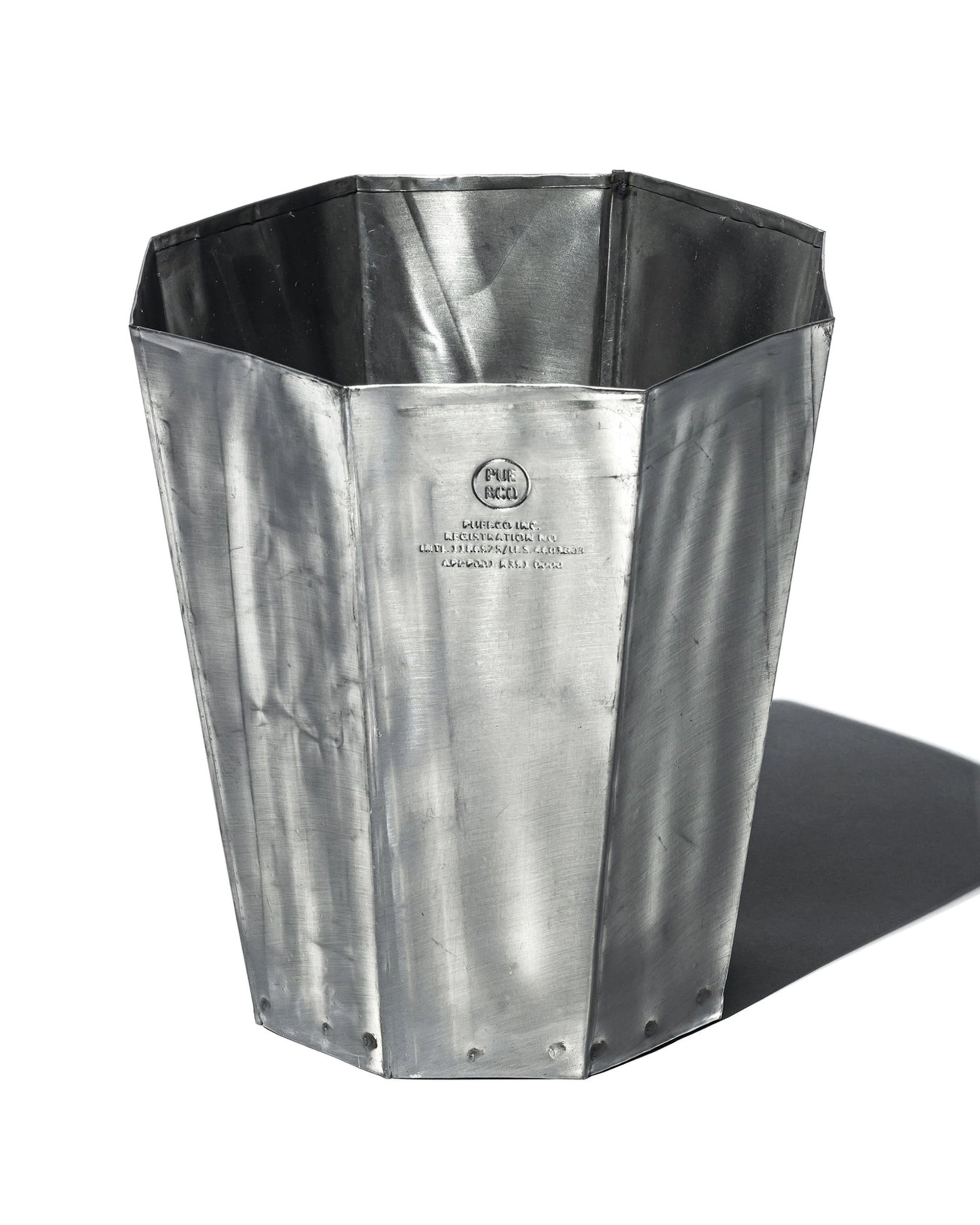 The Faire Steel Trashcan is a tall, octagonal recycled steel container with a flared rim, riveted base, and embossed logo and text on the front, evoking vintage oil cans and casting a shadow on a white background.
