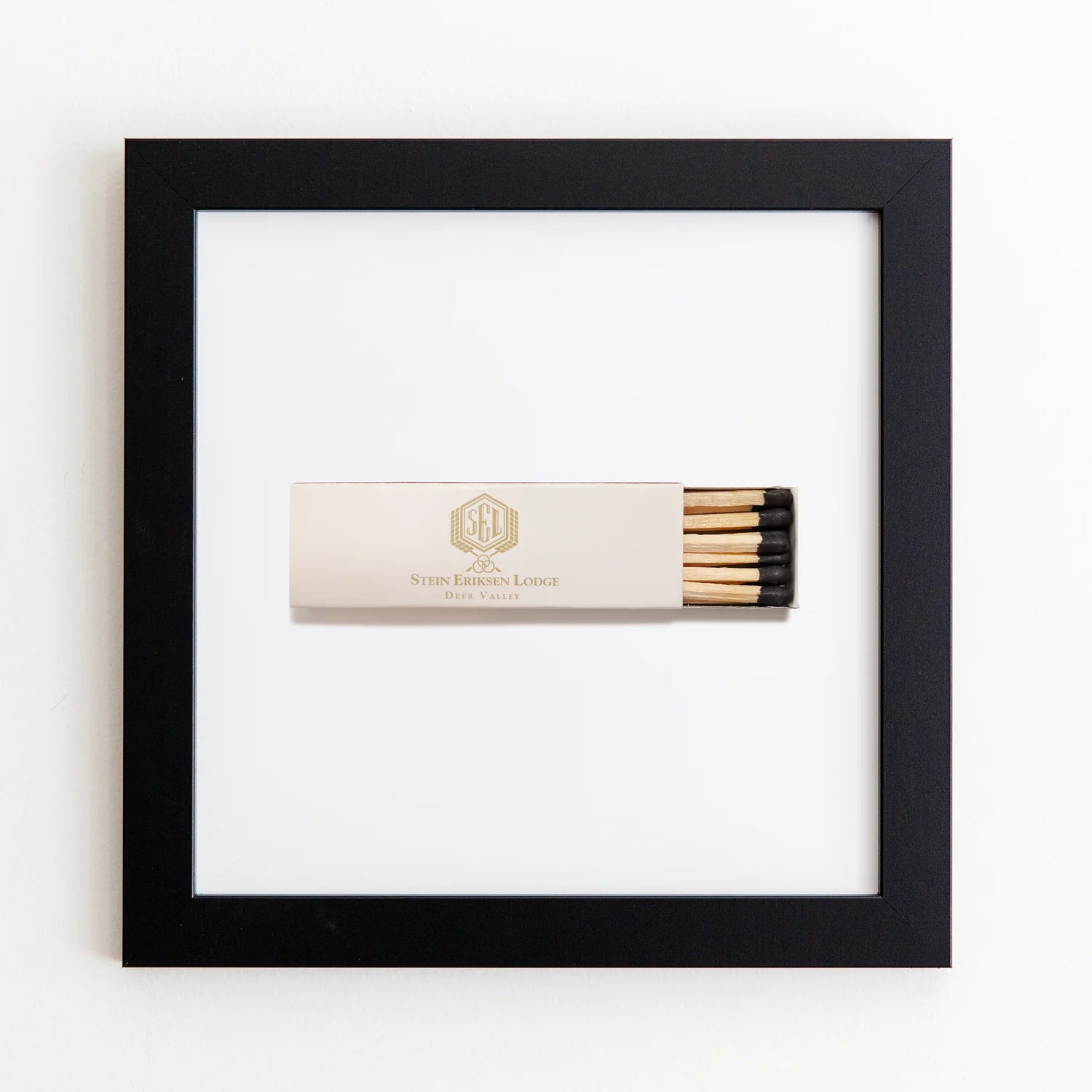A matchbox labeled &quot;Stein Eriksen Lodge Deer Valley&quot; is centered within a Art Square Black Frame against a white background. The matchbox is beige with a gold design and contains black-tipped matches.