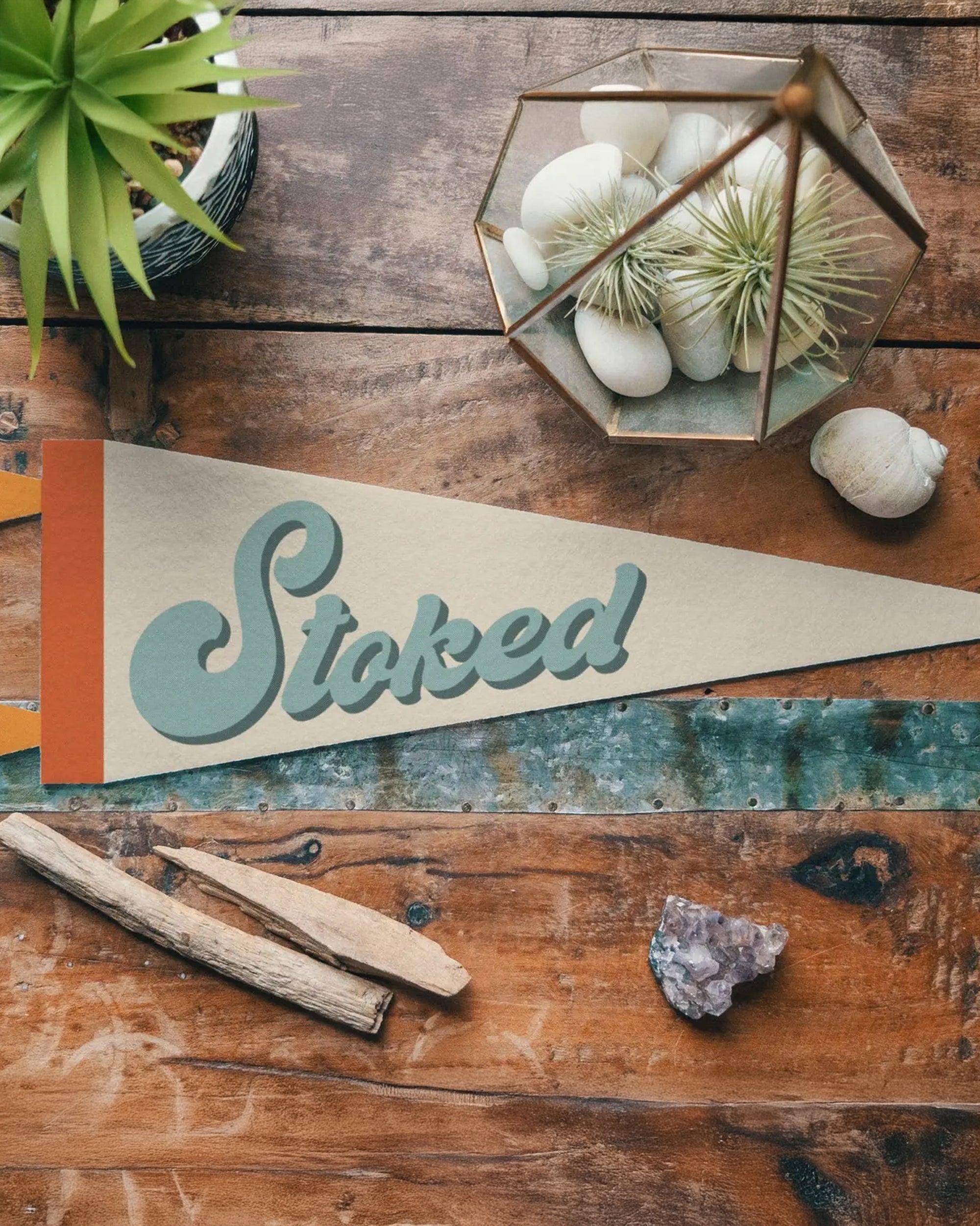 The Faire "Stoked" Felt Pennant rests on a rustic wooden table with crystals, driftwood, white stones, and potted plants—a perfect vintage-inspired surf decor accent for any room.