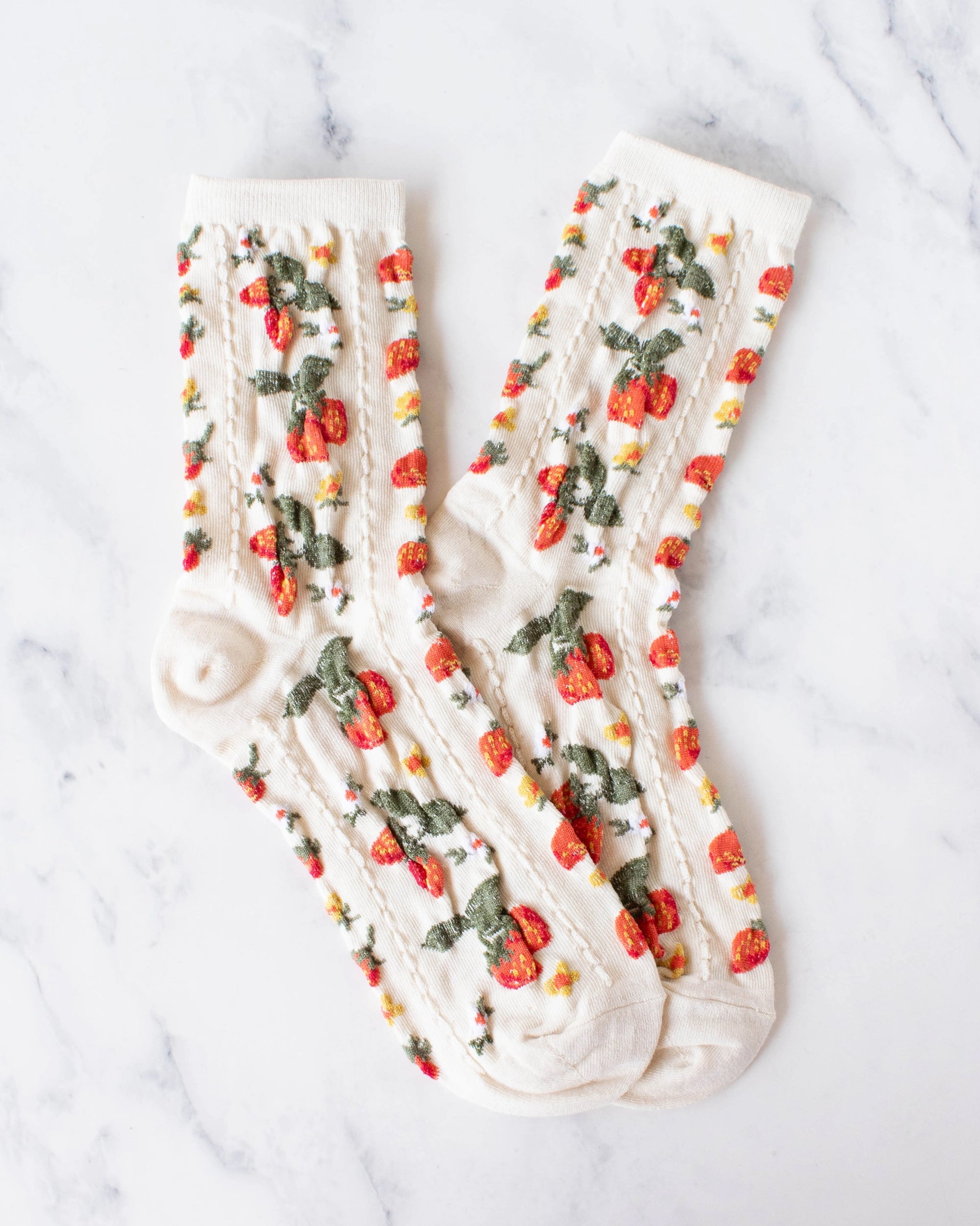 A pair of Faire Strawberry Farm Casual Socks in cream, featuring a strawberry and floral pattern, are crossed on white marble, bringing vintage charm to any casual outfit.