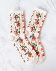 A pair of Faire Strawberry Farm Casual Socks in cream, featuring a strawberry and floral pattern, are crossed on white marble, bringing vintage charm to any casual outfit.