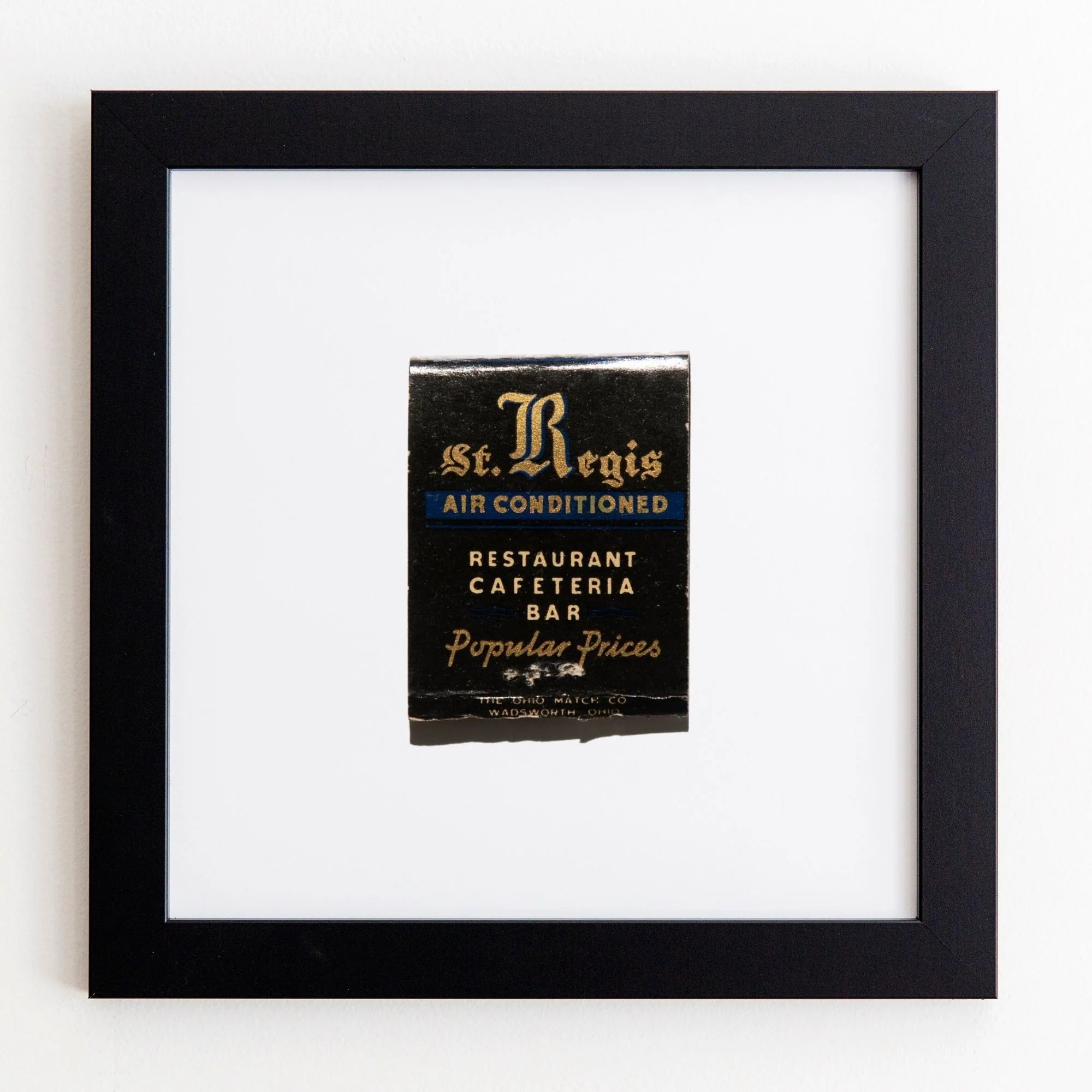A framed vintage advertisement for St. Regis Restaurant showing that it&#39;s air-conditioned, with offerings like a cafeteria and bar at popular prices, displayed in an Art Square Black Frame by Match South on an acrylic white wall.