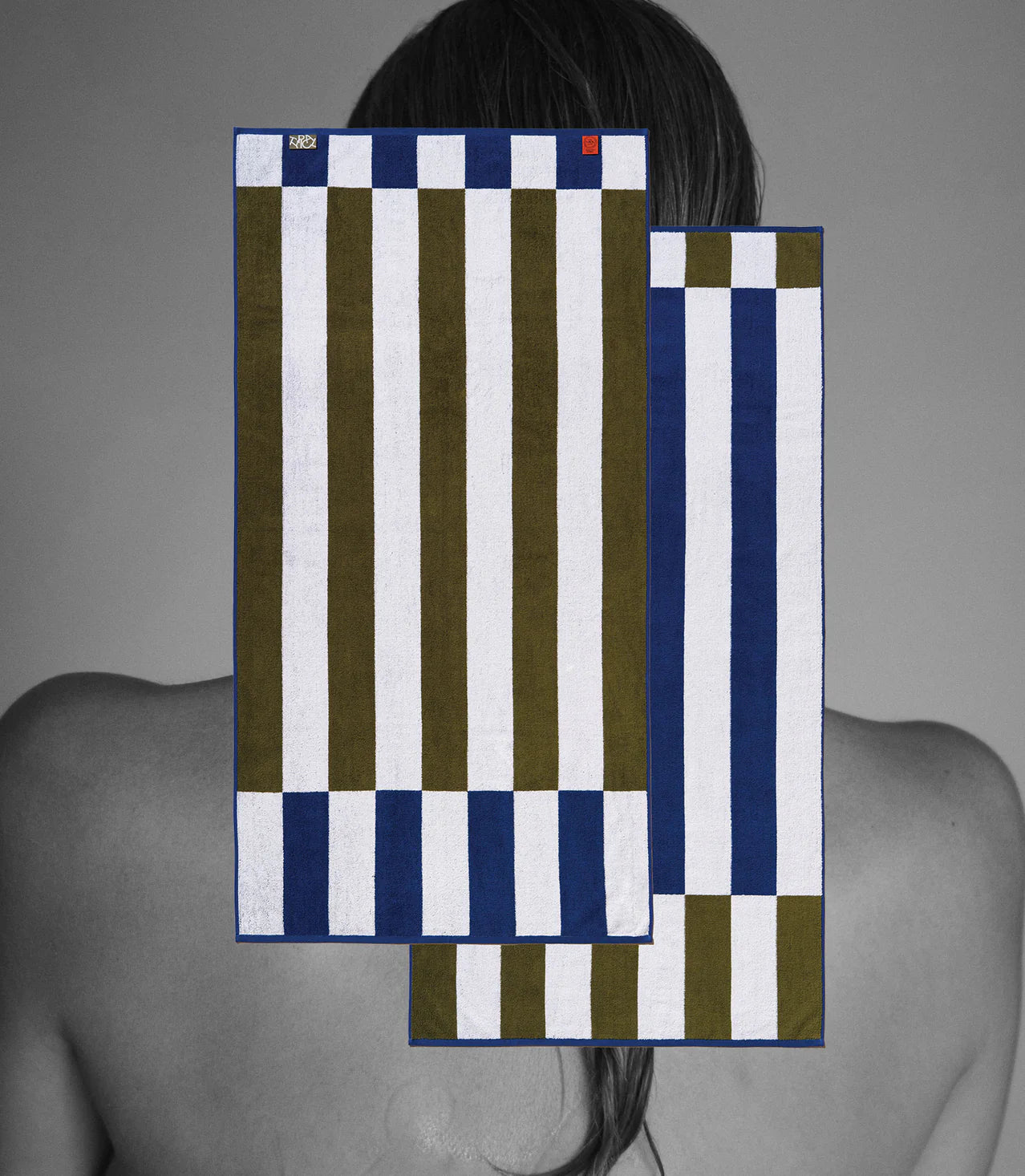 A grayscale photo of a person’s bare back and shoulders with two Typical Navy/Moss Stripe Bath Towels digitally superimposed in blue, olive green, and white over their upper back and hair.