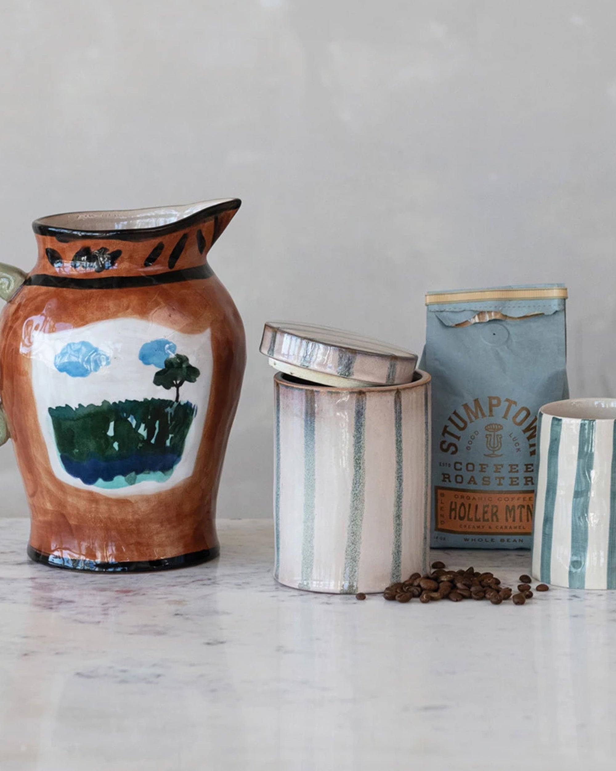 A hand-painted landscape jar, two Creative Co-op Striped Jars with Lid (one open), a blue Stumptown coffee bag, and scattered coffee beans rest on a marble surface.