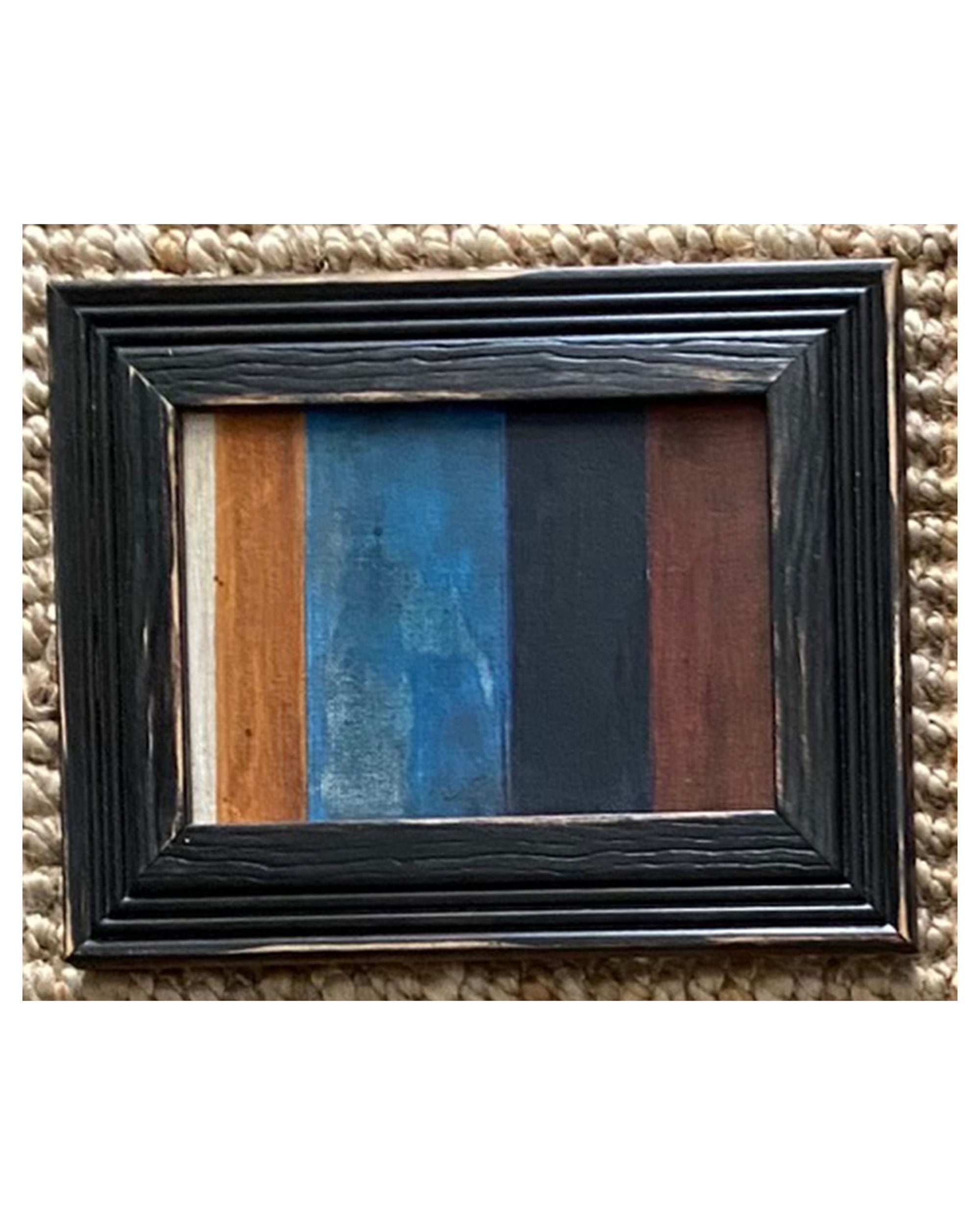 The STRIPES Framed Art by Tak Design is a small artwork featuring vertical stripes in cream, brown, blue, black, and dark brown hues, displayed on a textured beige woven surface.