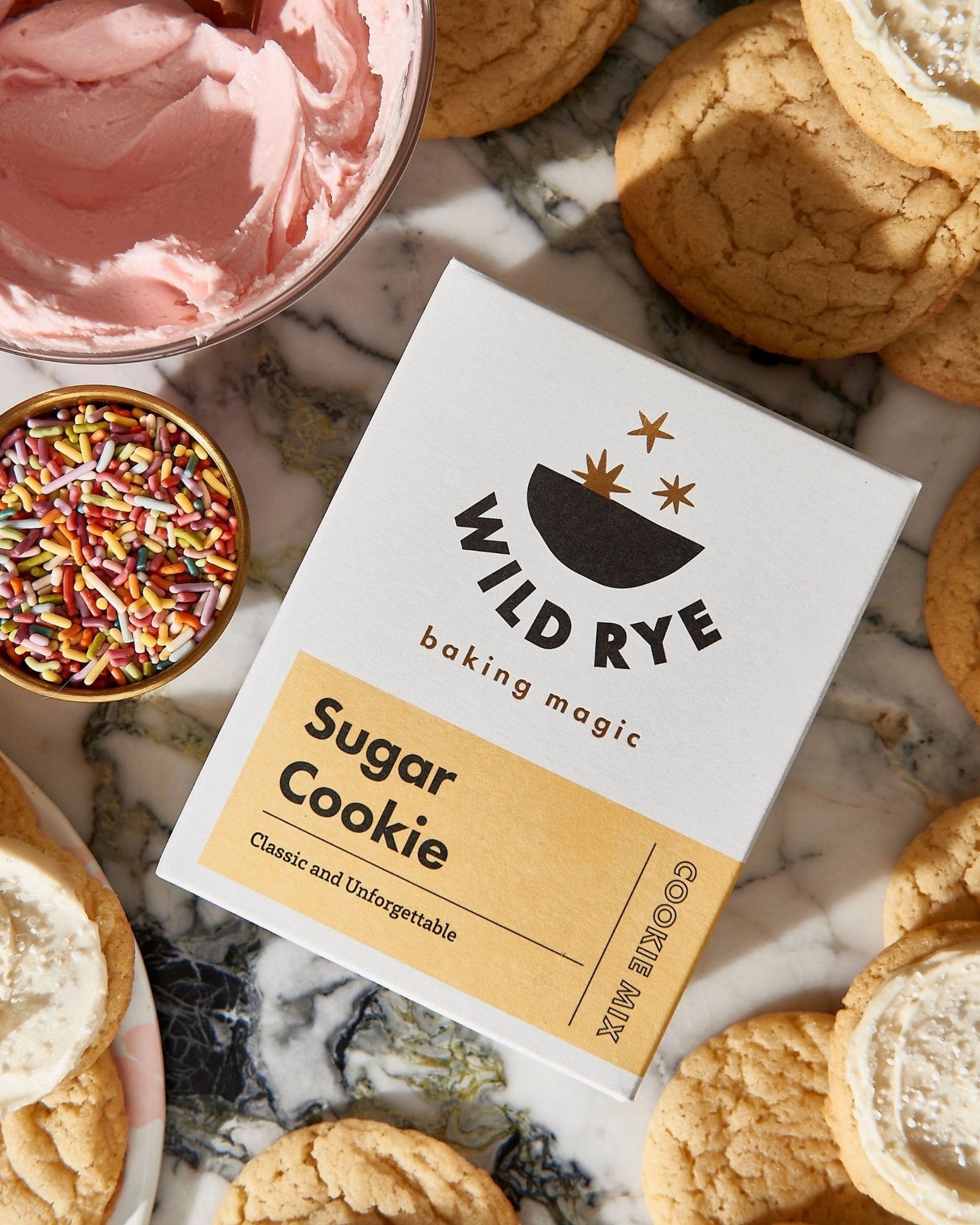 A box of Wild Rye Sugar Cookie Mix sits on a marble surface, surrounded by buttery cookies, pink frosting, a bowl of sprinkles, and a few vanilla cookies topped with frosting.