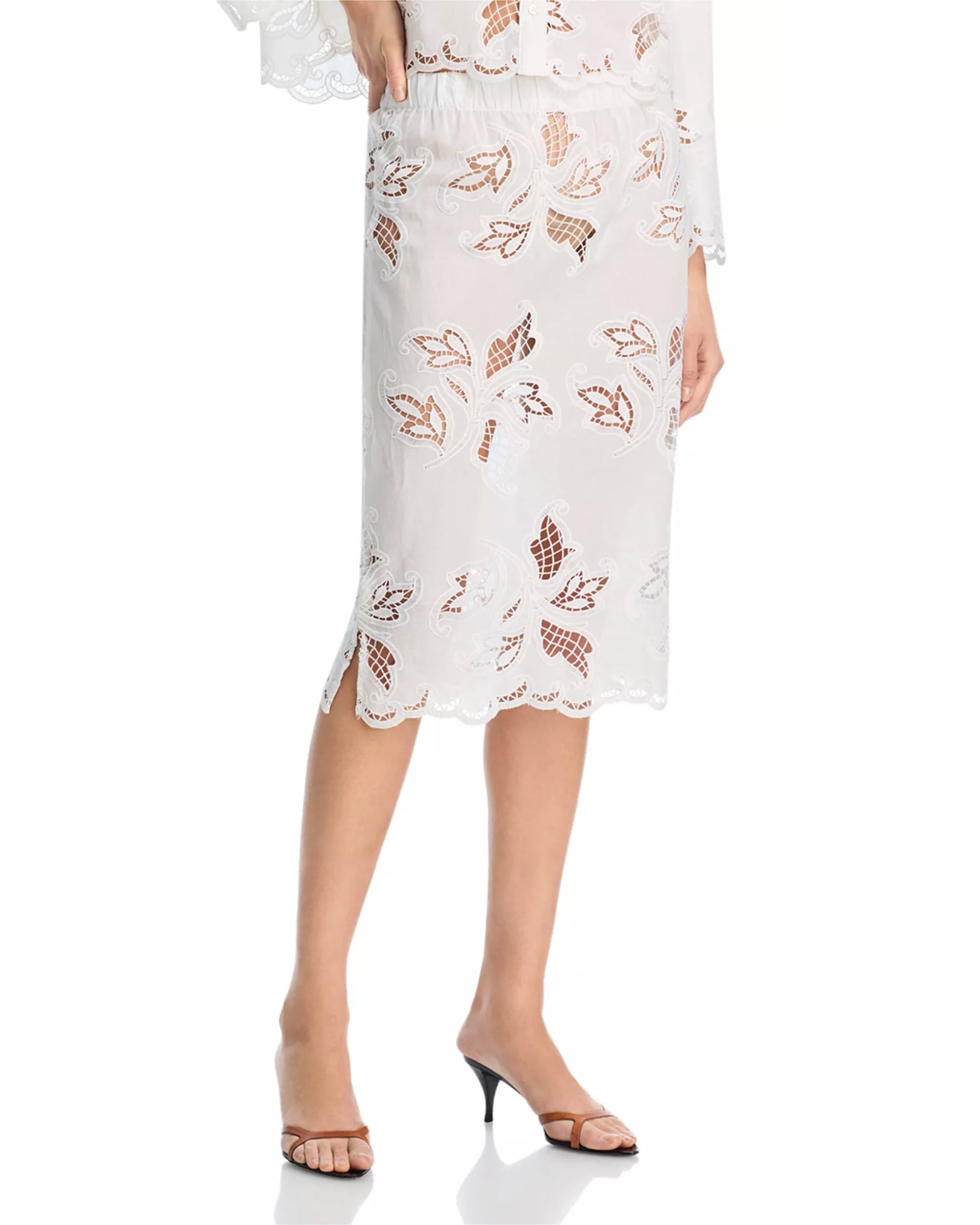 A woman’s lower half is shown wearing the Cissa Summer Pencil Skirt—white, knee-length with floral cutout lace and scalloped edges—paired with open-toe heeled sandals.
