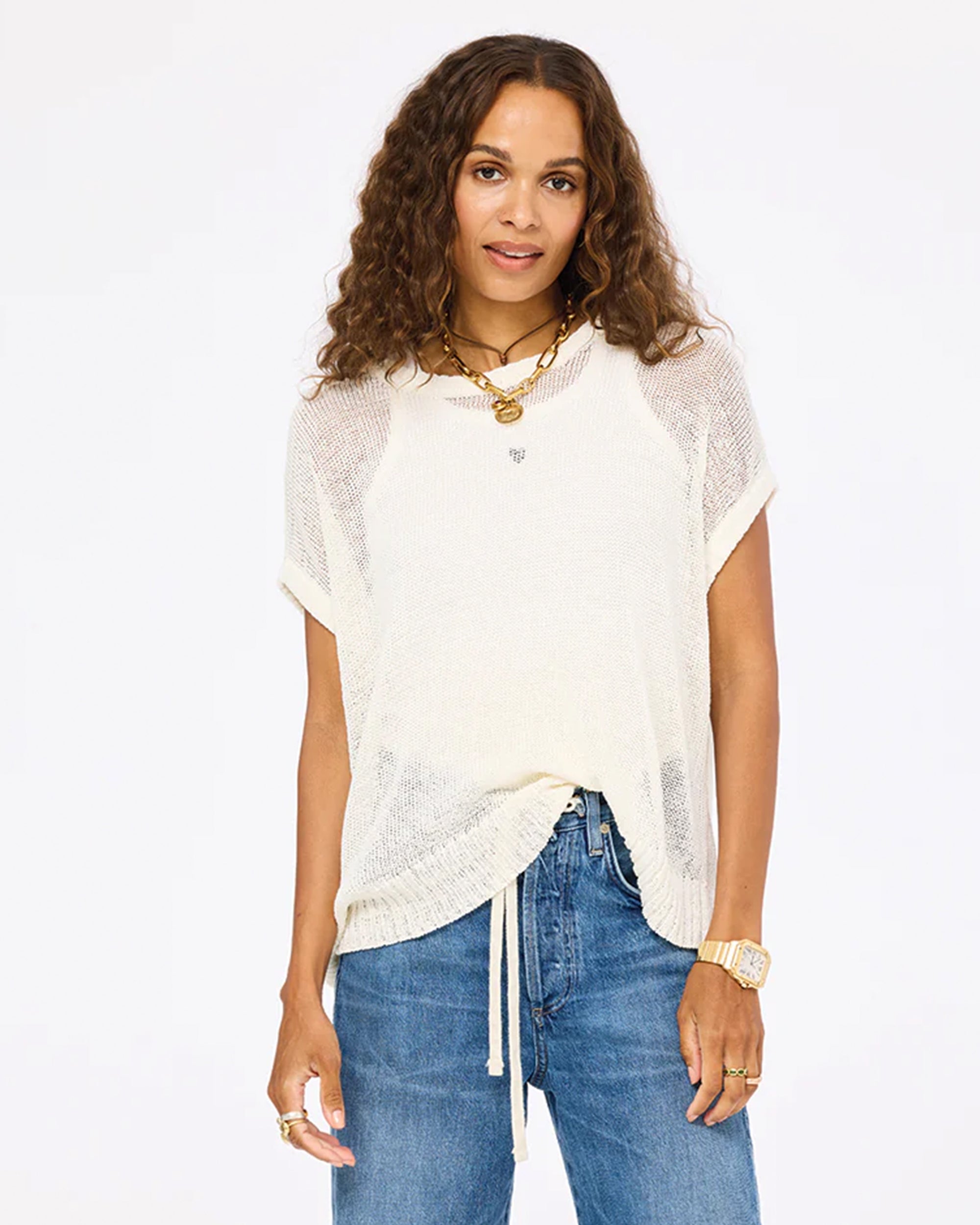 A woman with long curly hair wears the Kerri Rosenthal Summer Vestie Sweater over a tank and blue jeans, accessorized with layered necklaces, a watch, and rings, standing against a plain white background.