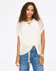 A woman with long curly hair wears the Kerri Rosenthal Summer Vestie Sweater over a tank and blue jeans, accessorized with layered necklaces, a watch, and rings, standing against a plain white background.