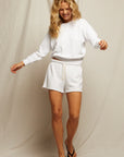 A woman with long blonde hair smiles while wearing a white sweatshirt and Perfectwhitetee Summer Loop Terry Sweat Shorts with an elastic waistband, paired with black flip-flops, standing against a light-colored fabric backdrop.