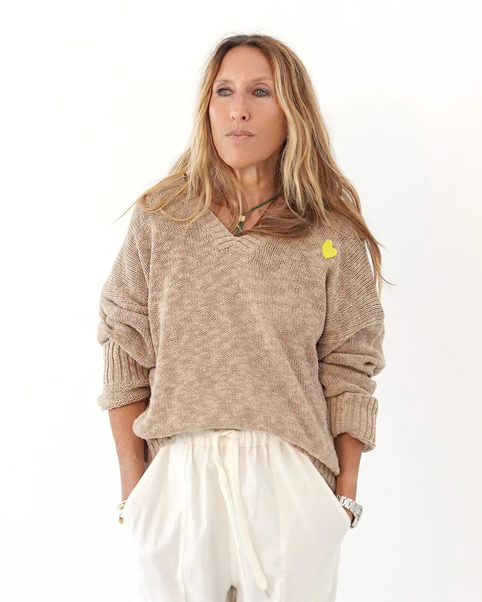 A woman with long, light brown hair wears the Kerri Rosenthal Summerland V Neck Sweater with sleeves rolled up and loose white pants, standing against a white background with her hands in her pockets.