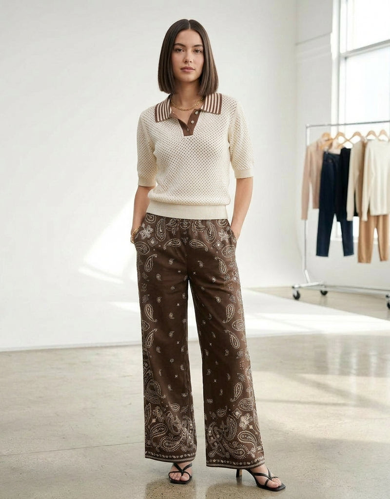 A woman with straight shoulder-length hair stands indoors, hands in her pockets, wearing SUNCOO's Woven Pantalon Jessy and a cream knit top with a striped collar, near hanging clothes on a polished floor.
