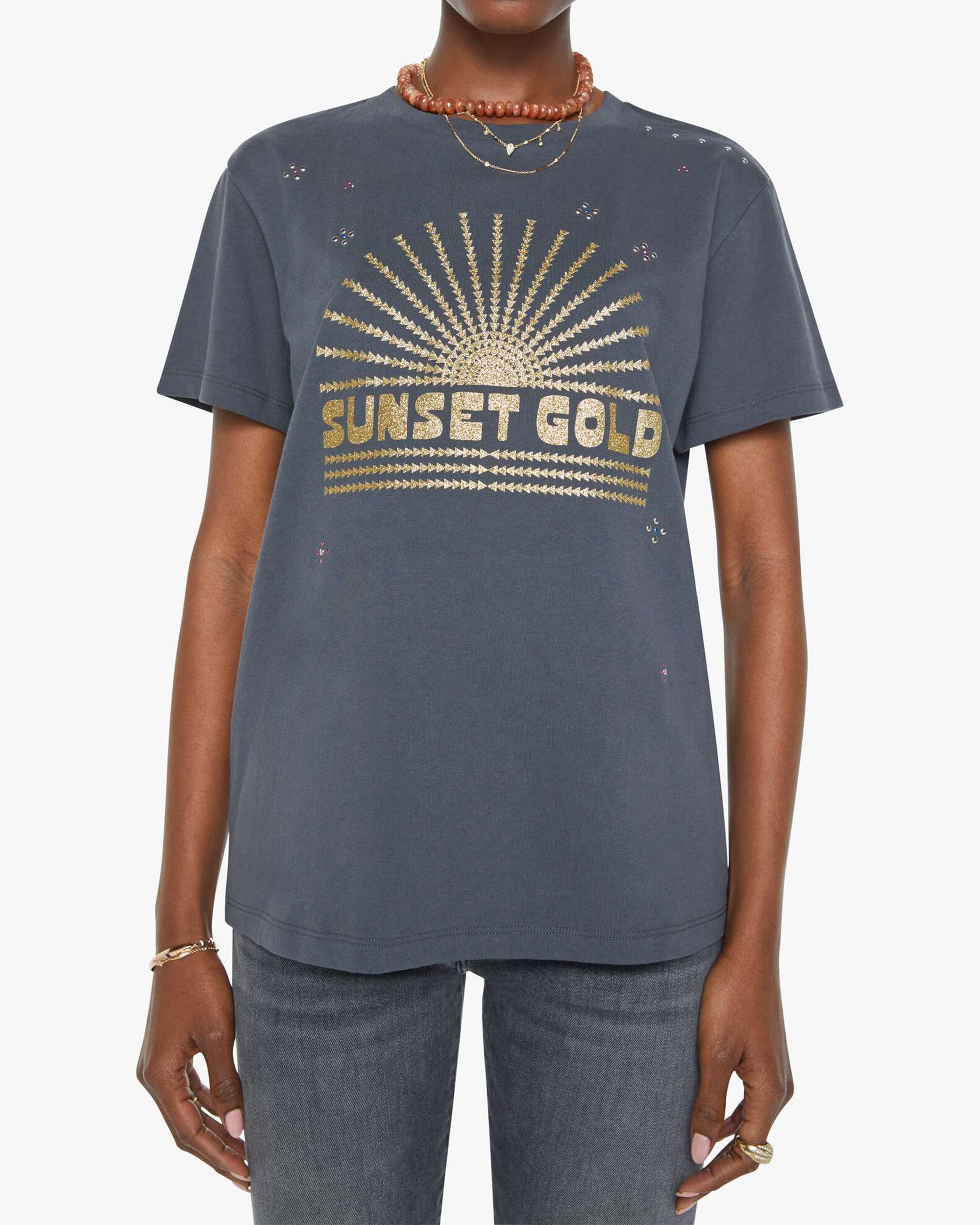 Someone wearing the Rowdy crewneck tee by Mother in dark gray with a gold sunburst and "SUNSET GOLD" on the front, styled oversized with blue jeans, layered necklaces, and bracelets. Made from 100% cotton for comfort.