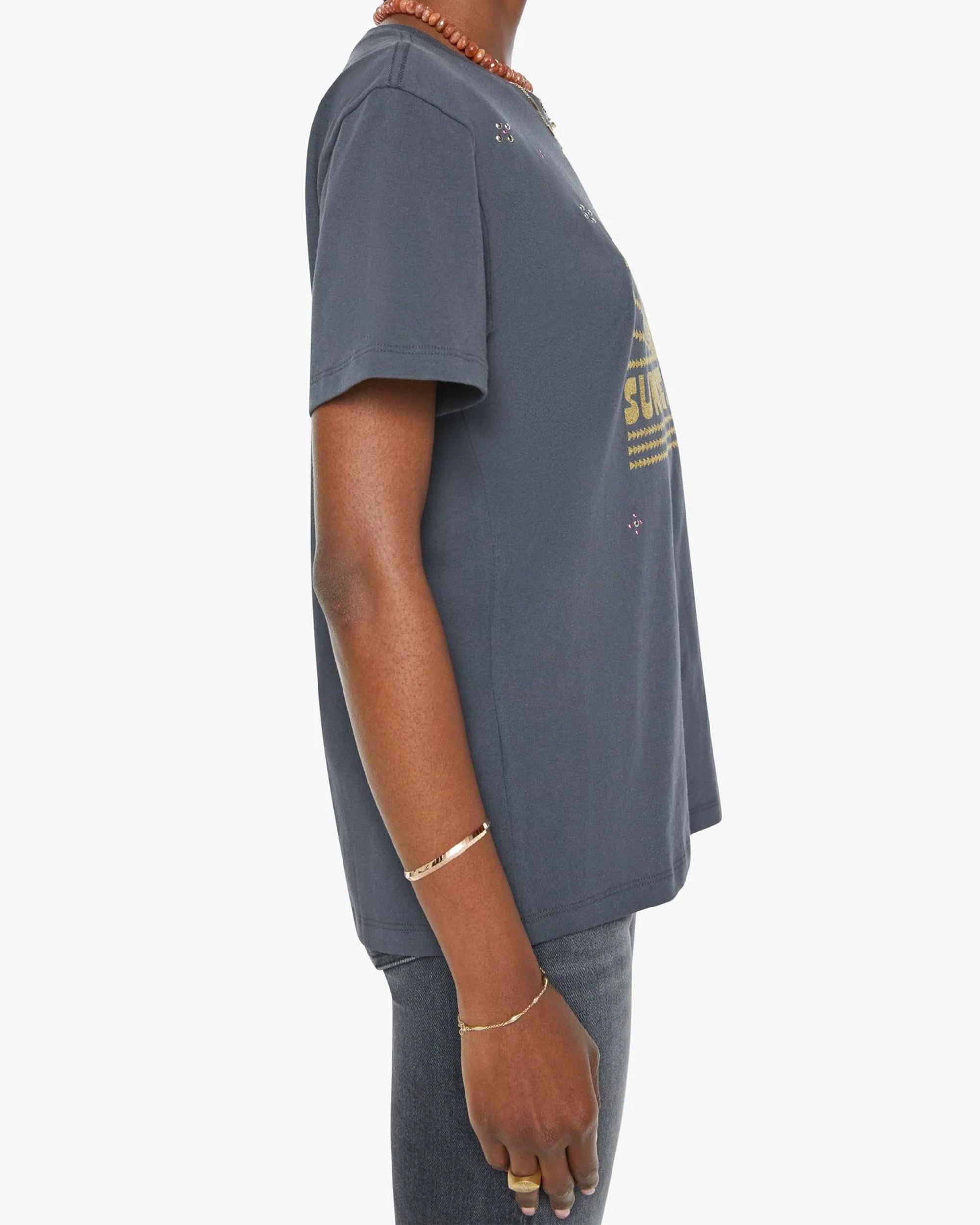A person wearing the Rowdy, a navy blue 100% cotton crewneck tee by Mother, paired with dark jeans and gold wrist and finger jewelry, shown from the side against a plain white background.