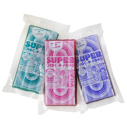 Three colorful Super Choc-O-Food chocolate bars by Faire, in green, pink, and purple retro-style wrappers, rest in clear sleeves against a white background. These darkened milk chocolate treats feature collectible packaging that attracts enthusiasts.