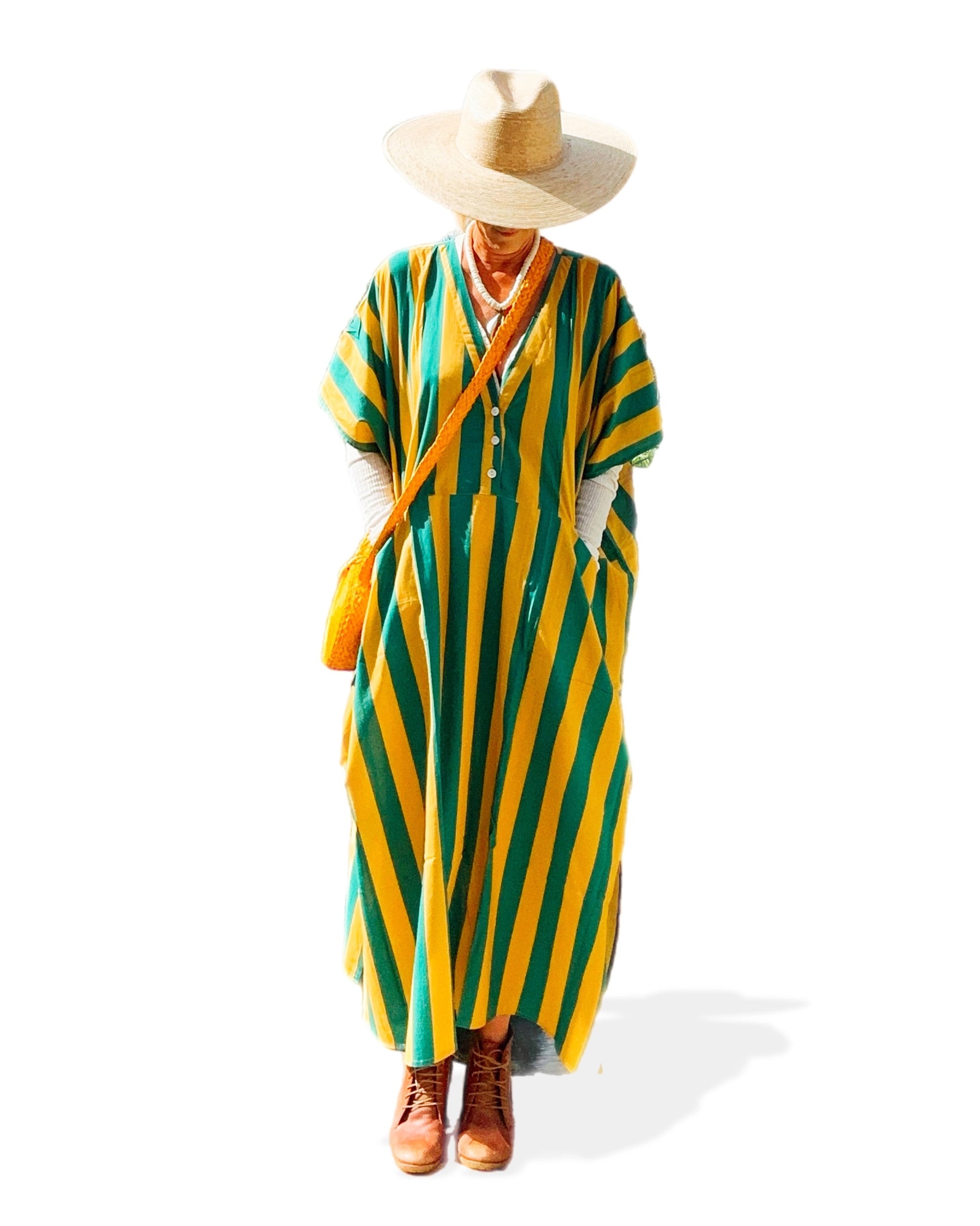 A person wears the Faire Suft Stripe Kaftan—a lightweight cotton kaftan with green and yellow stripes—plus a wide-brimmed straw hat, brown boots, and a yellow bag, standing against a white background.