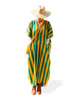 A person wears the Faire Suft Stripe Kaftan—a lightweight cotton kaftan with green and yellow stripes—plus a wide-brimmed straw hat, brown boots, and a yellow bag, standing against a white background.