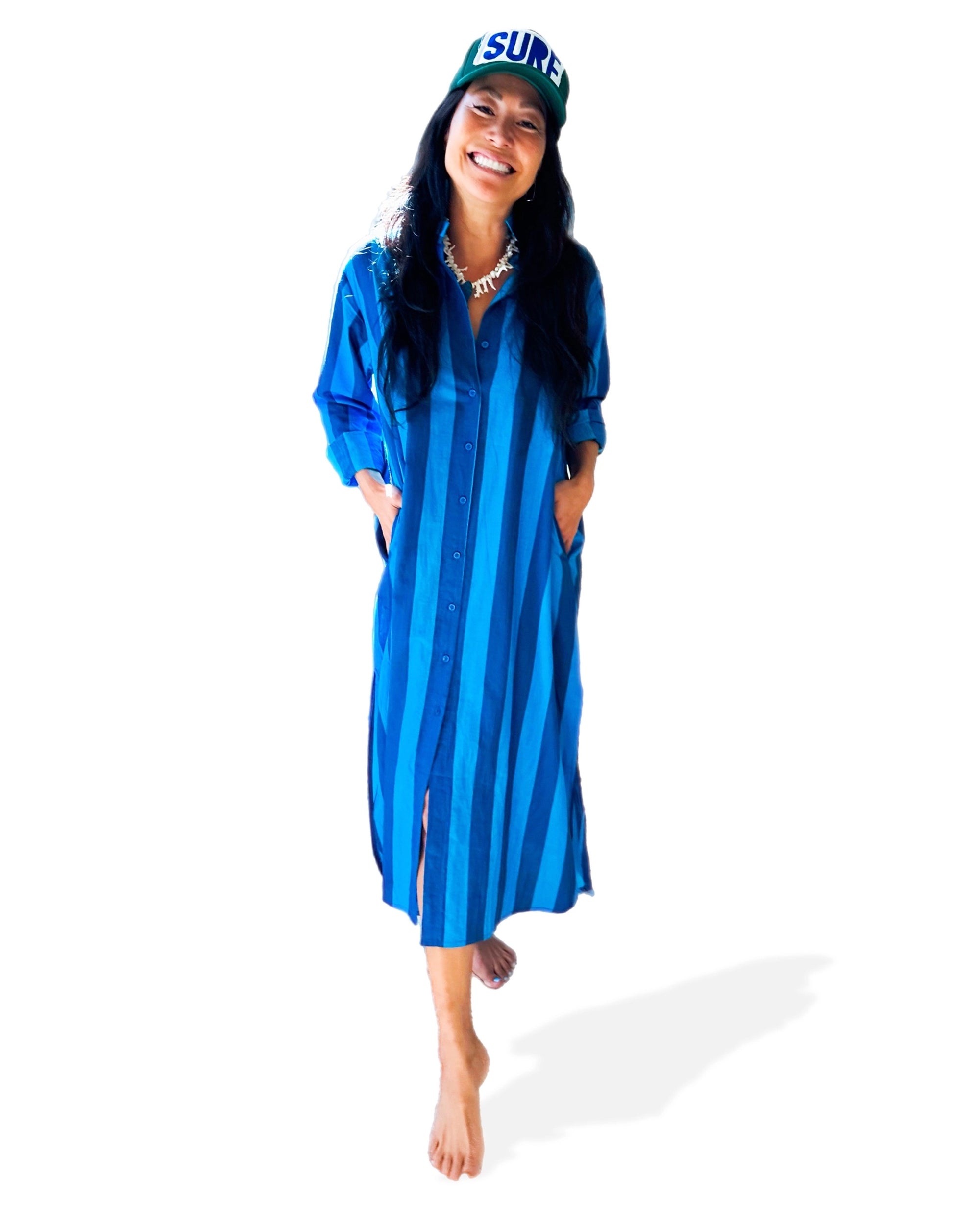 A smiling person, barefoot, wears the Faire Surf Stripe Shirt Dress—blue cotton with buttons—paired with a green-and-white "SURF" cap. They stand hands-in-pockets against a white background.
