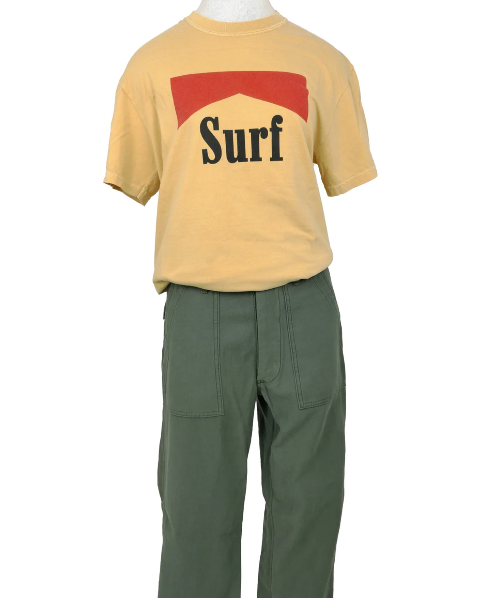 A mannequin models the SURF Tshirt by Hype Factory Inc.—a beige, vintage-washed, 100% cotton tee with a red graphic and "Surf" in black. Features unisex sizing for comfort, paired here with high-waisted green pants.