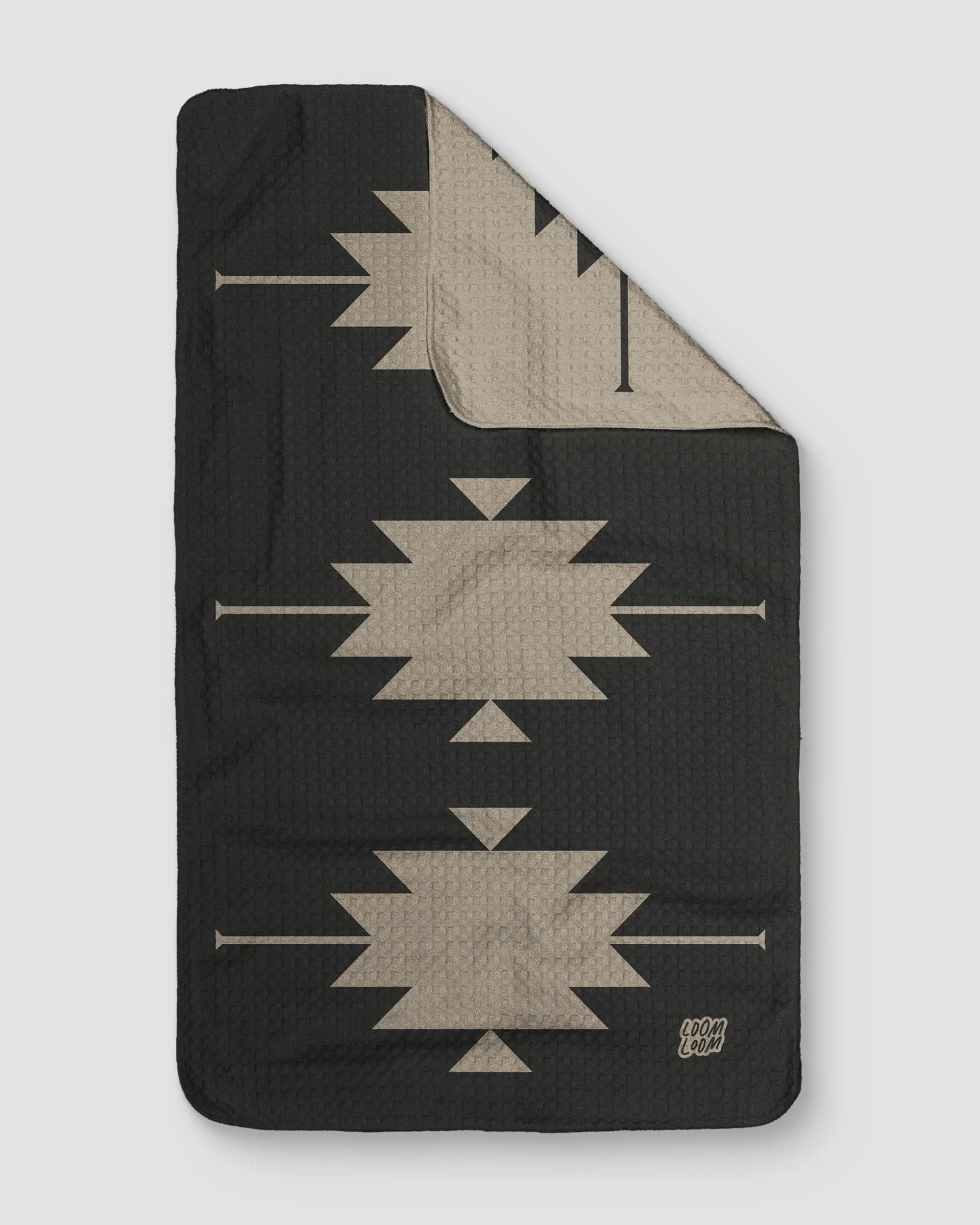 The SW Gray Towel by Faire is a rectangular, quilted towel made from EcoLoom fabric with a geometric Southwestern pattern in beige on black. One corner is folded to reveal the reverse side with inverted colors.