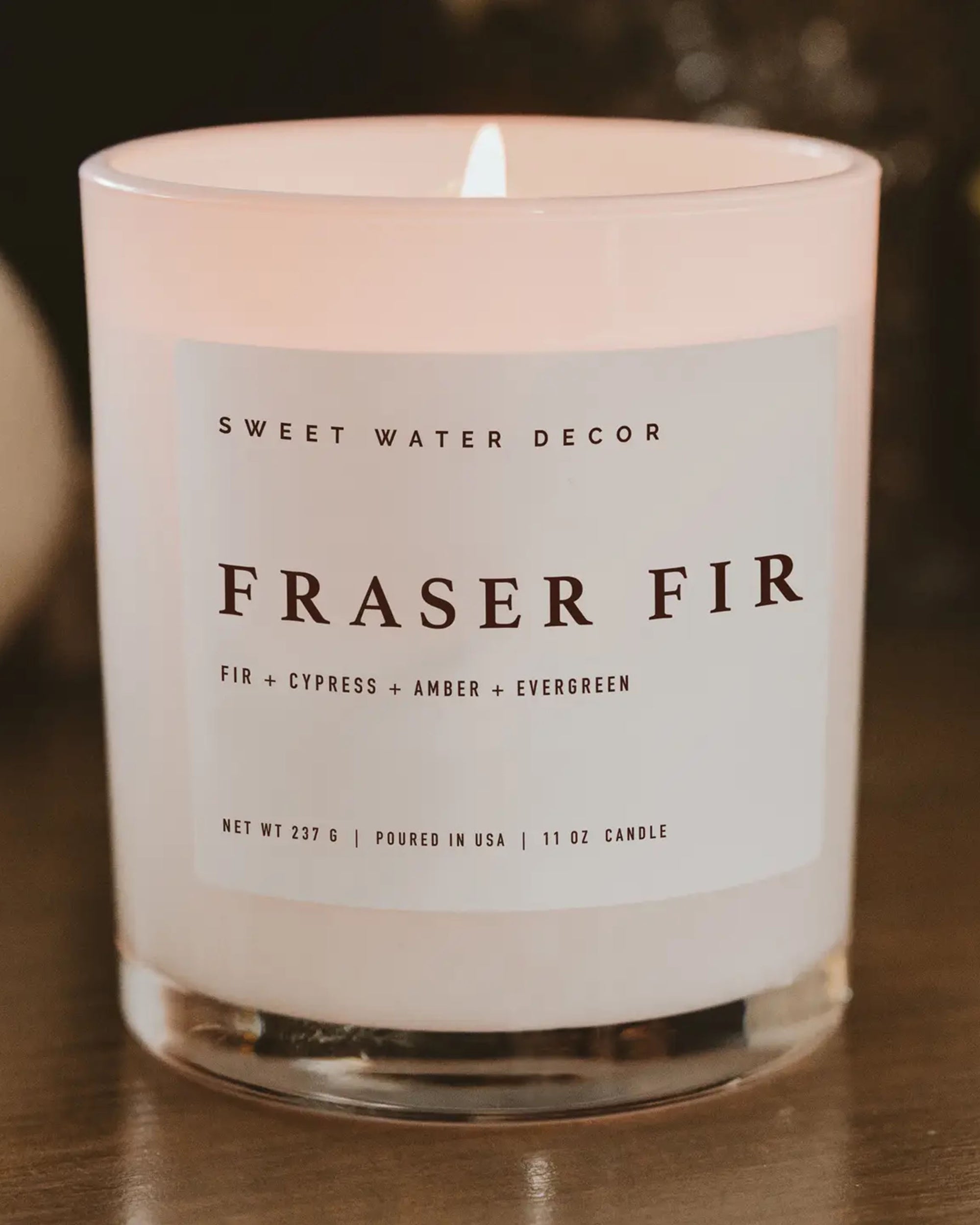 A lit Faire Christmas Candle w/ Lid, a white soy-based candle in a clear glass jar labeled "Fraser Fir, Fir + Cypress + Amber + Evergreen," made with premium fragrance oil, sits on wood for a long burn time.