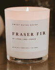 A lit Faire Christmas Candle w/ Lid, a white soy-based candle in a clear glass jar labeled "Fraser Fir, Fir + Cypress + Amber + Evergreen," made with premium fragrance oil, sits on wood for a long burn time.