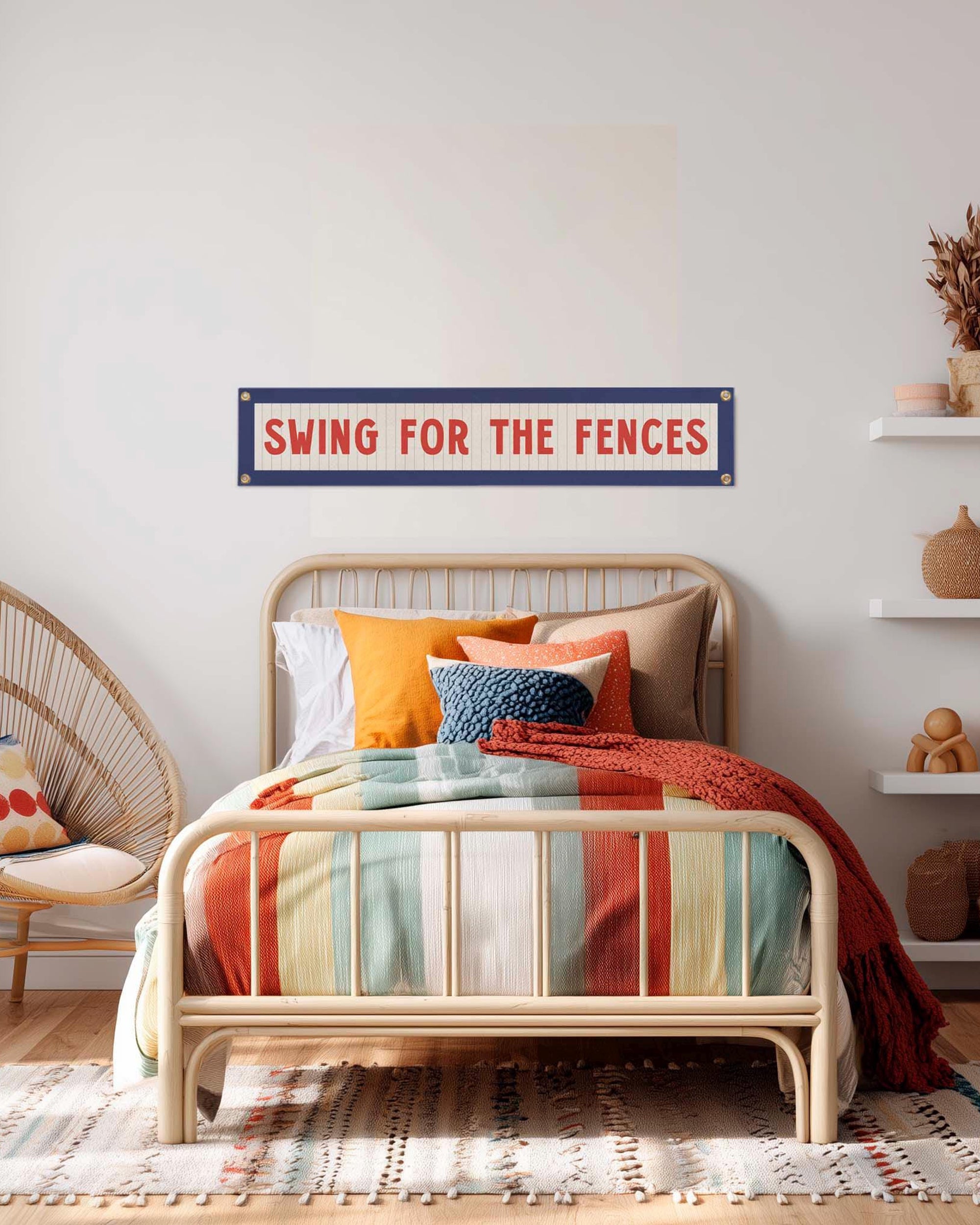 A cozy bedroom features a rattan bed with colorful striped bedding and orange accent pillows. Above, the Faire Swing for the Fences Banner adds vintage charm. Shelves with decor and a rattan chair complete the look.