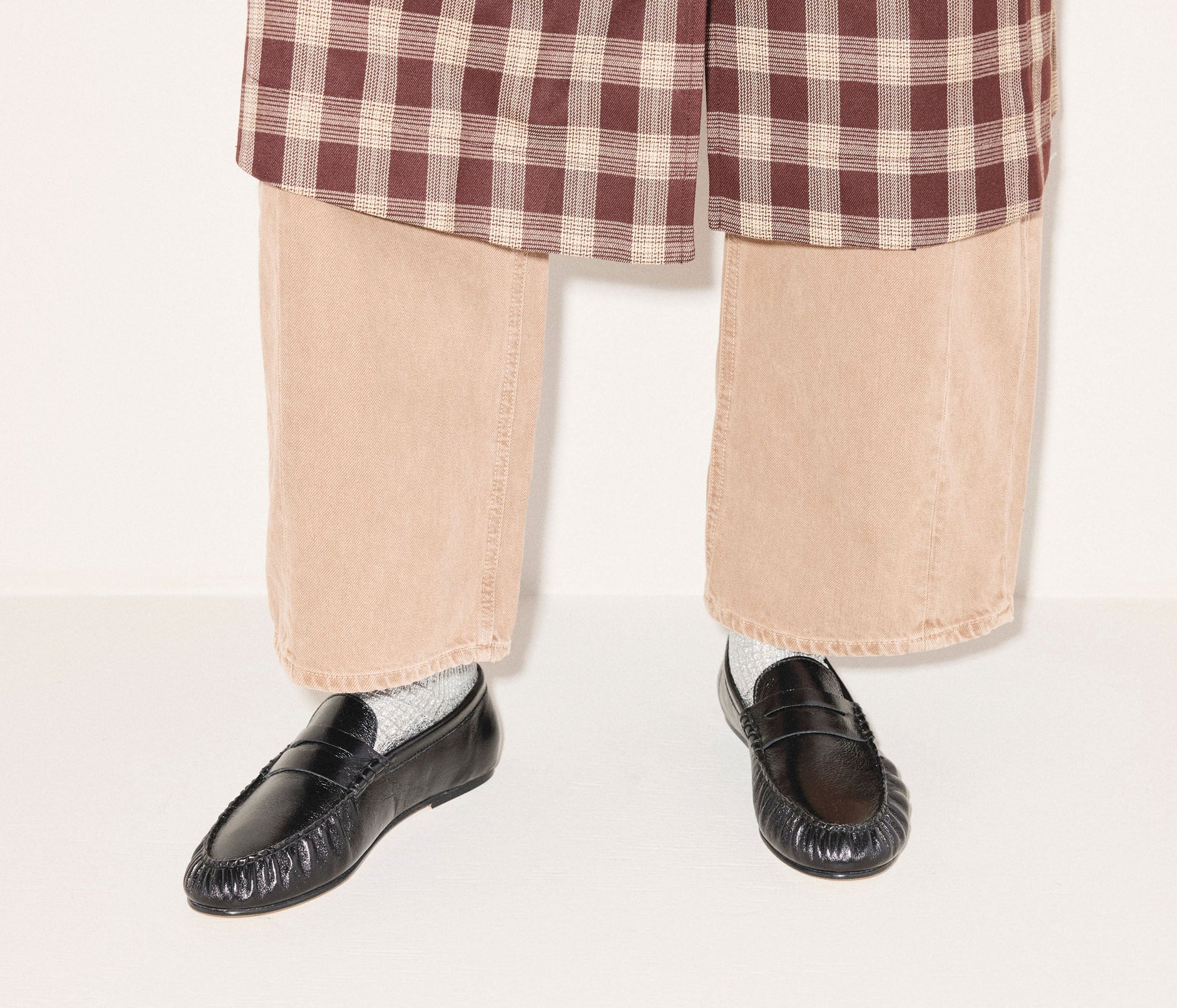 A person stands against a plain light background wearing beige wide-legged pants, a red and white plaid coat, gray socks, and black Sydney Crinkle Loafers by Freda Salvador.