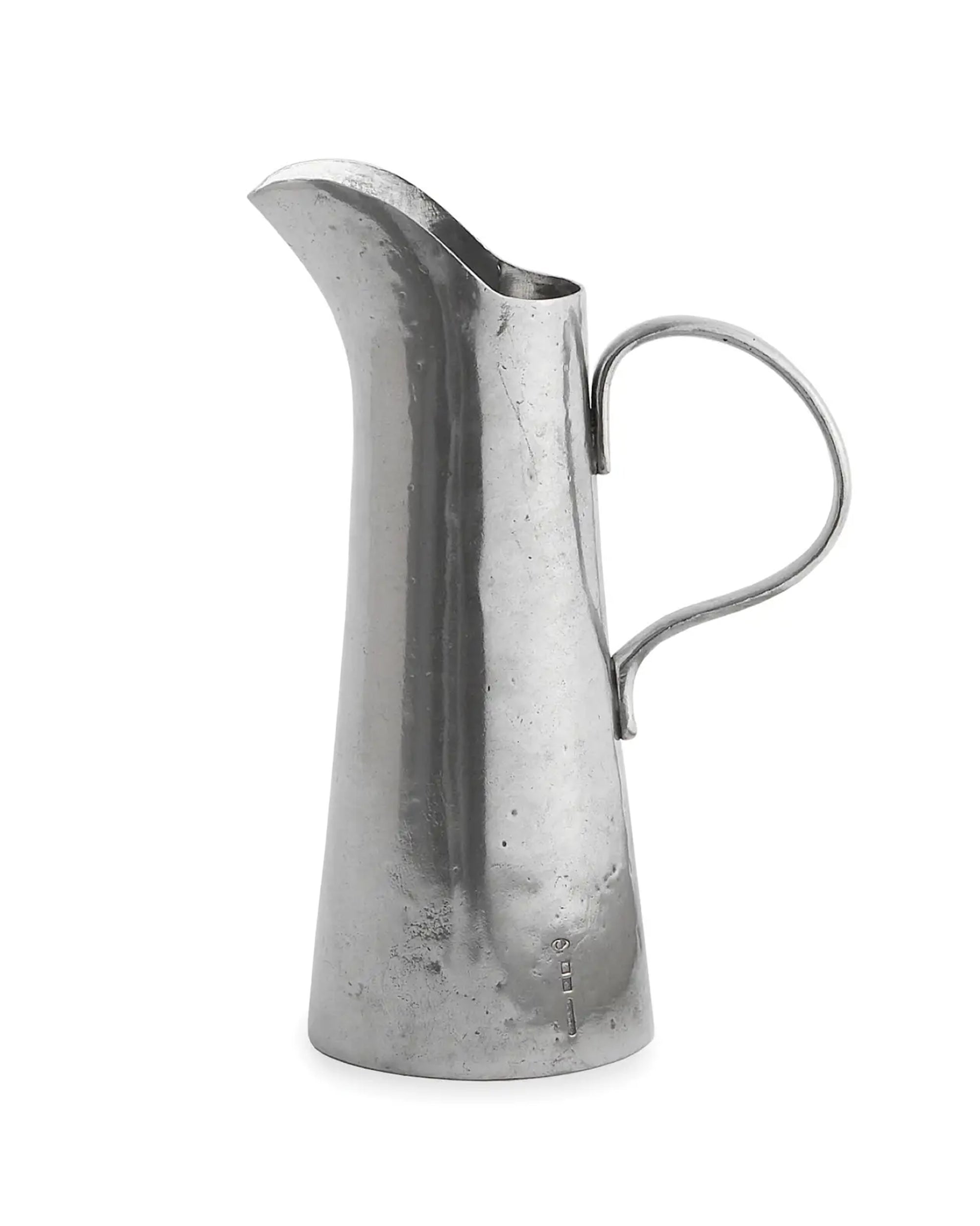The VIN2270 Vintage Tall Tapered Pitcher by Faire is a tall, narrow silver pitcher with a curved handle and wide spout, hand made in Italy from artisan pewter with a subtly textured, smooth finish.